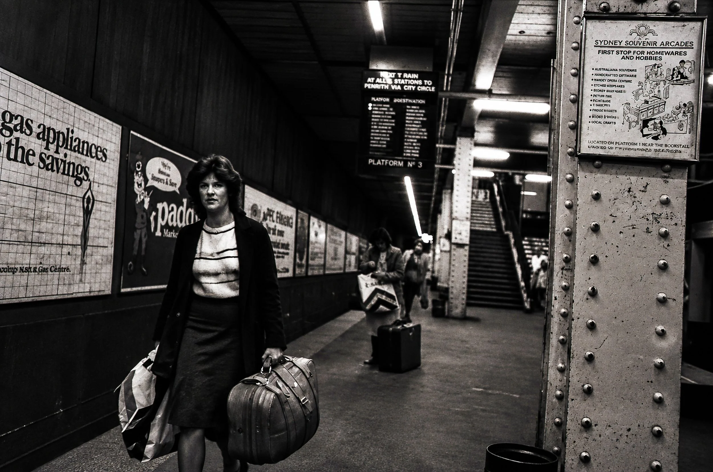 Through the Underground - A lone traveller walks along the platform carrying luggage while other passengers disappear into the distance. The repeating advertisements, lights and structural columns emphasise the depth of the underground station.