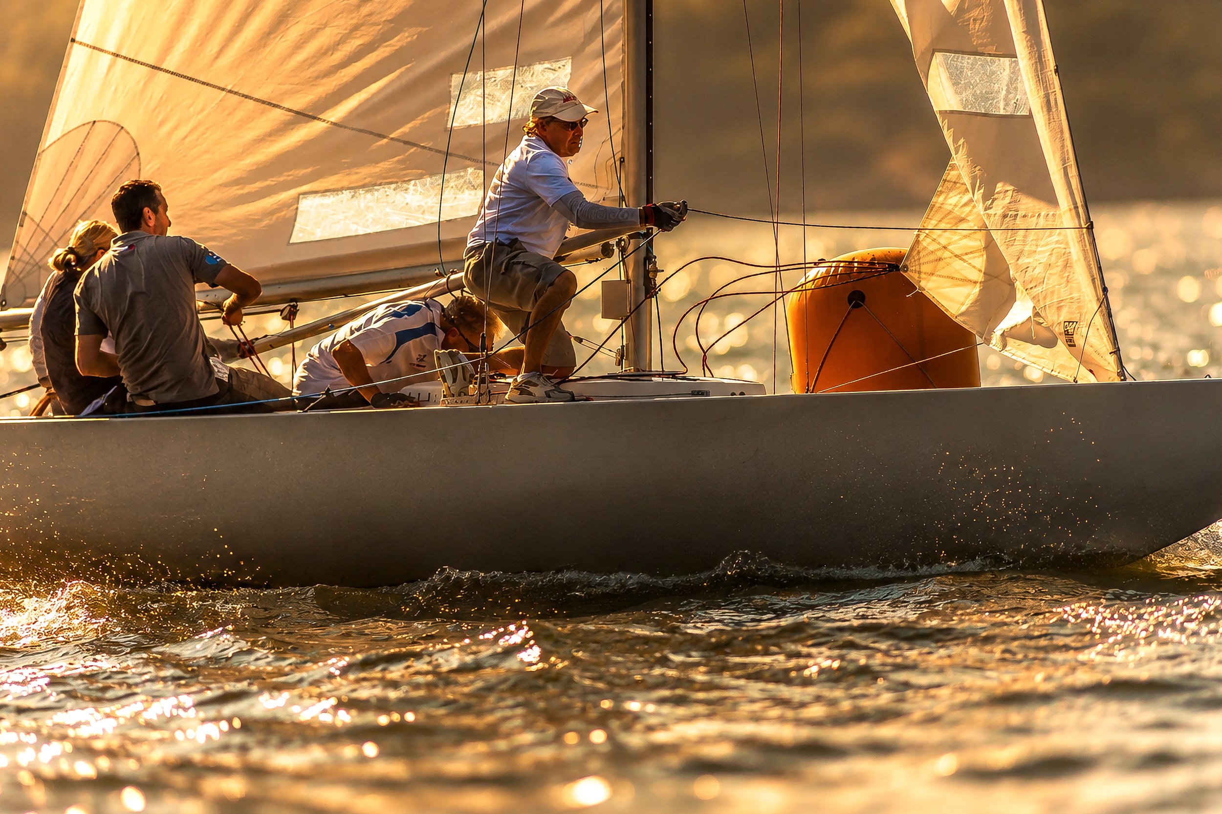A sailboat with four crew members sailing during sunset or sunrise, with golden light reflecting off the water.