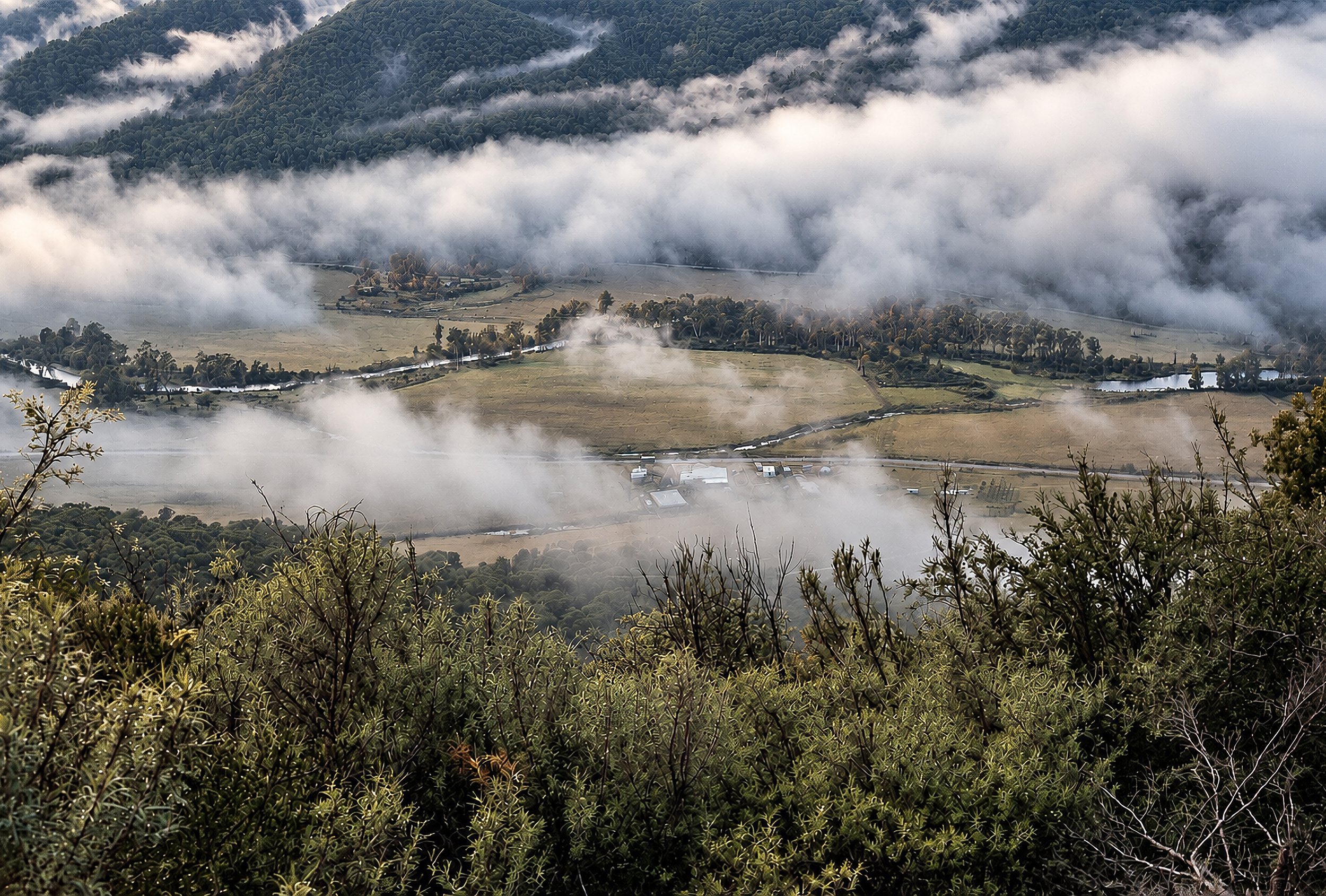 Valley of Mist - A broad mountain valley emerges through broken cloud, with fields, waterways and scattered trees appearing and disappearing beneath the mist. The subtle colour brings out the greens and muted earth tones of the Australian landscape w