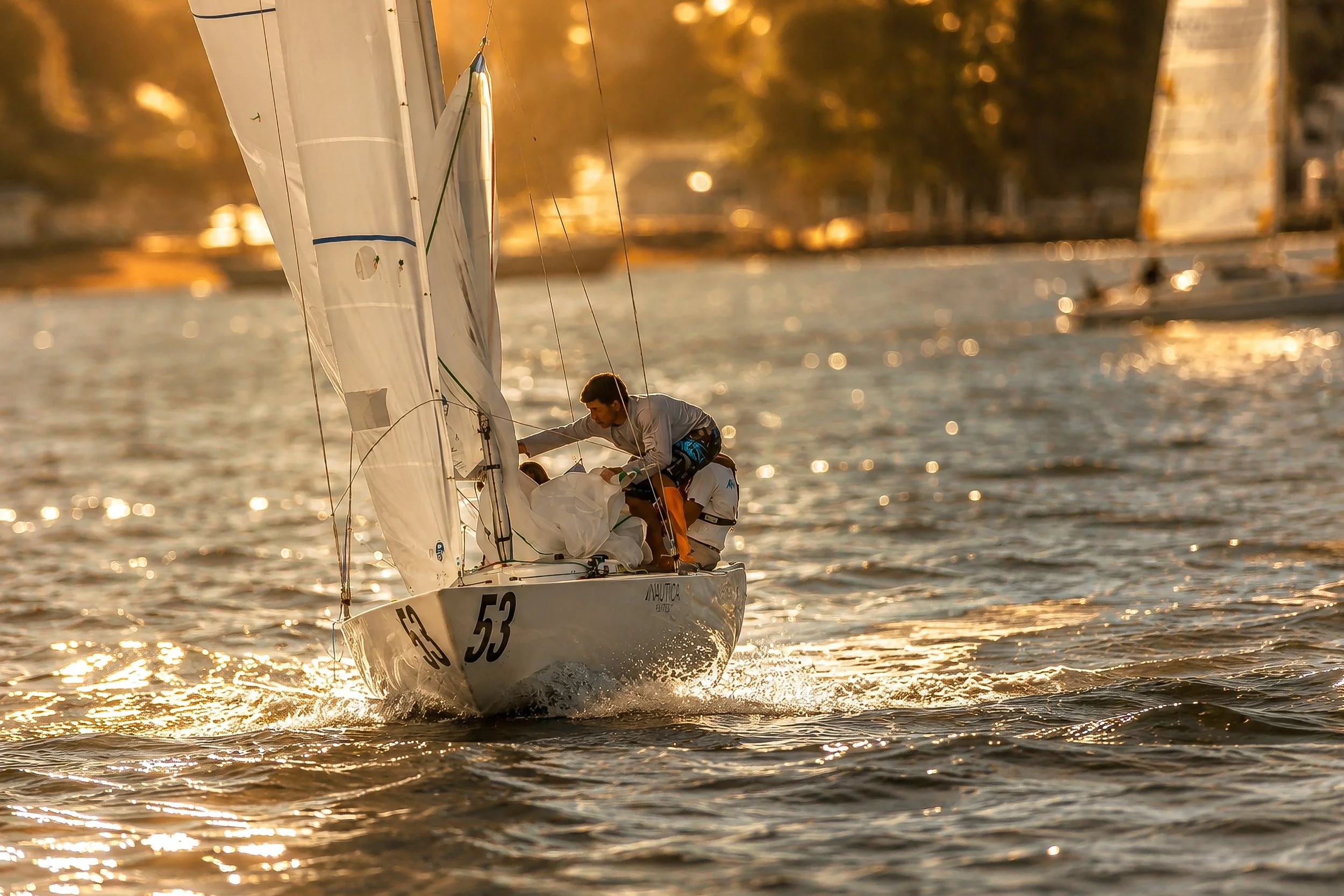 A sailboat with the number 53 on its side, sailing on a body of water during sunset. The boat has two sailors onboard adjusting the sails. The scene is warm and golden, with reflections on the water and blurred background trees.
