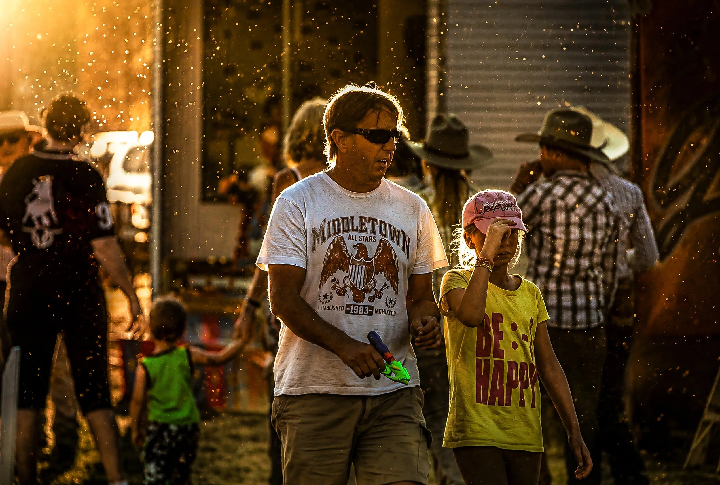 A man and a girl walking outdoors during sunset, surrounded by other people, some wearing hats, with specks of dust or pollen illuminated in the sunlight.