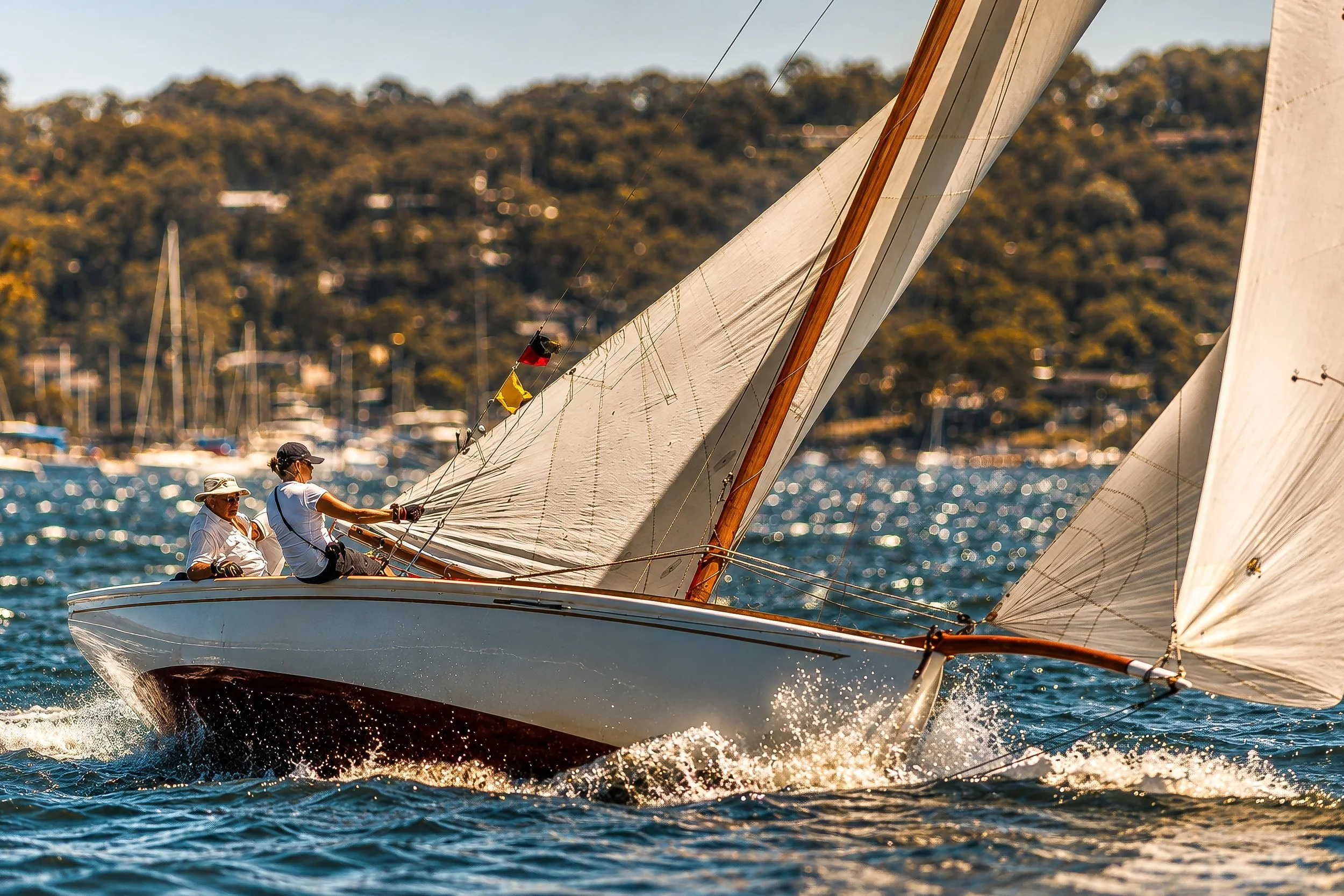 Sailboats racing on a body of water with a hilly, tree-lined shoreline in the background.