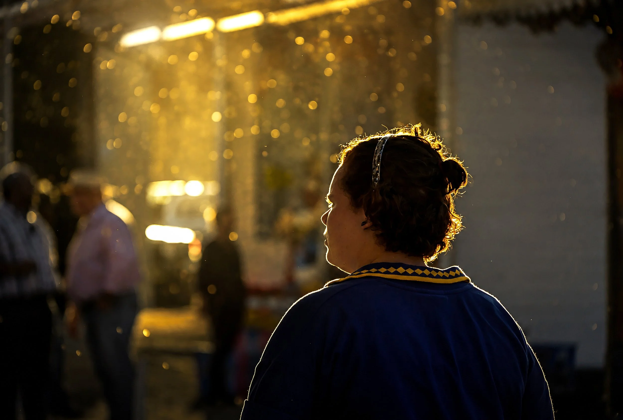 A woman with curly hair and a headband stands outdoors during sunset, with blurred figures and warm golden light in the background.