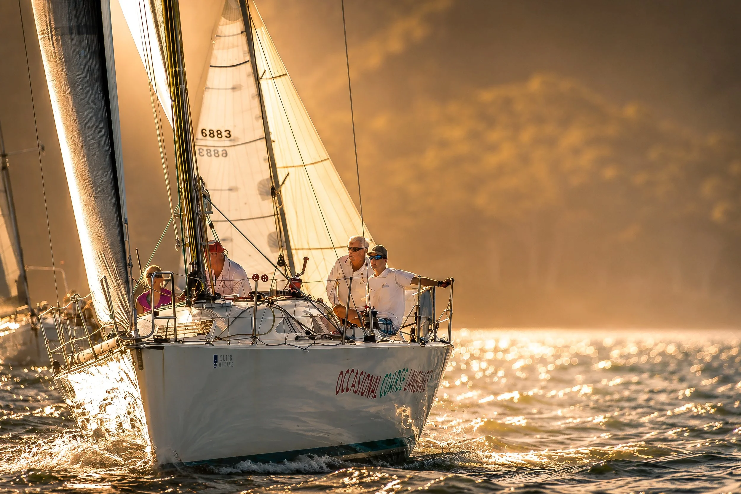 A sailboat with multiple people on deck sailing on the water during sunset. The boat is white with a large sail, and the sunlight reflects off the water's surface, creating a golden glow.