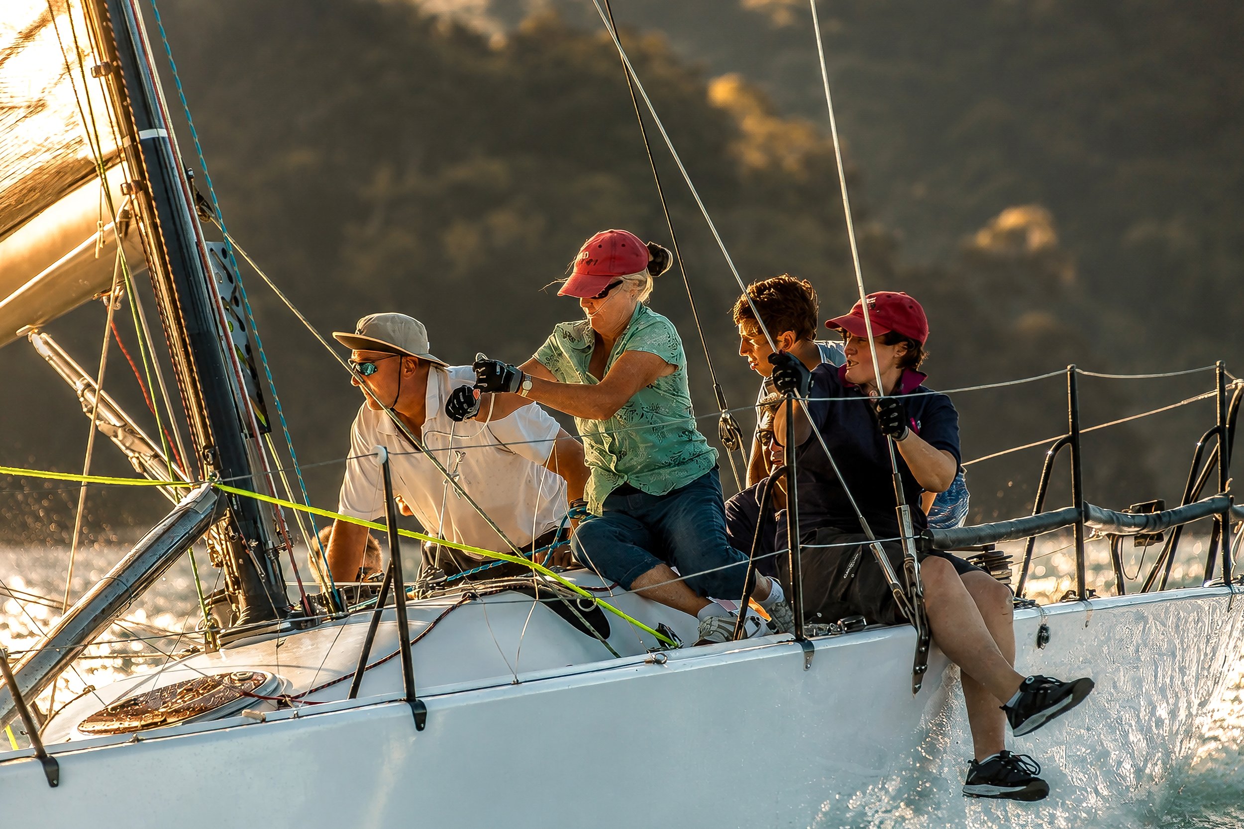 A group of people sailing on a boat during sunset, with some in red caps and gloves, working together on the deck.