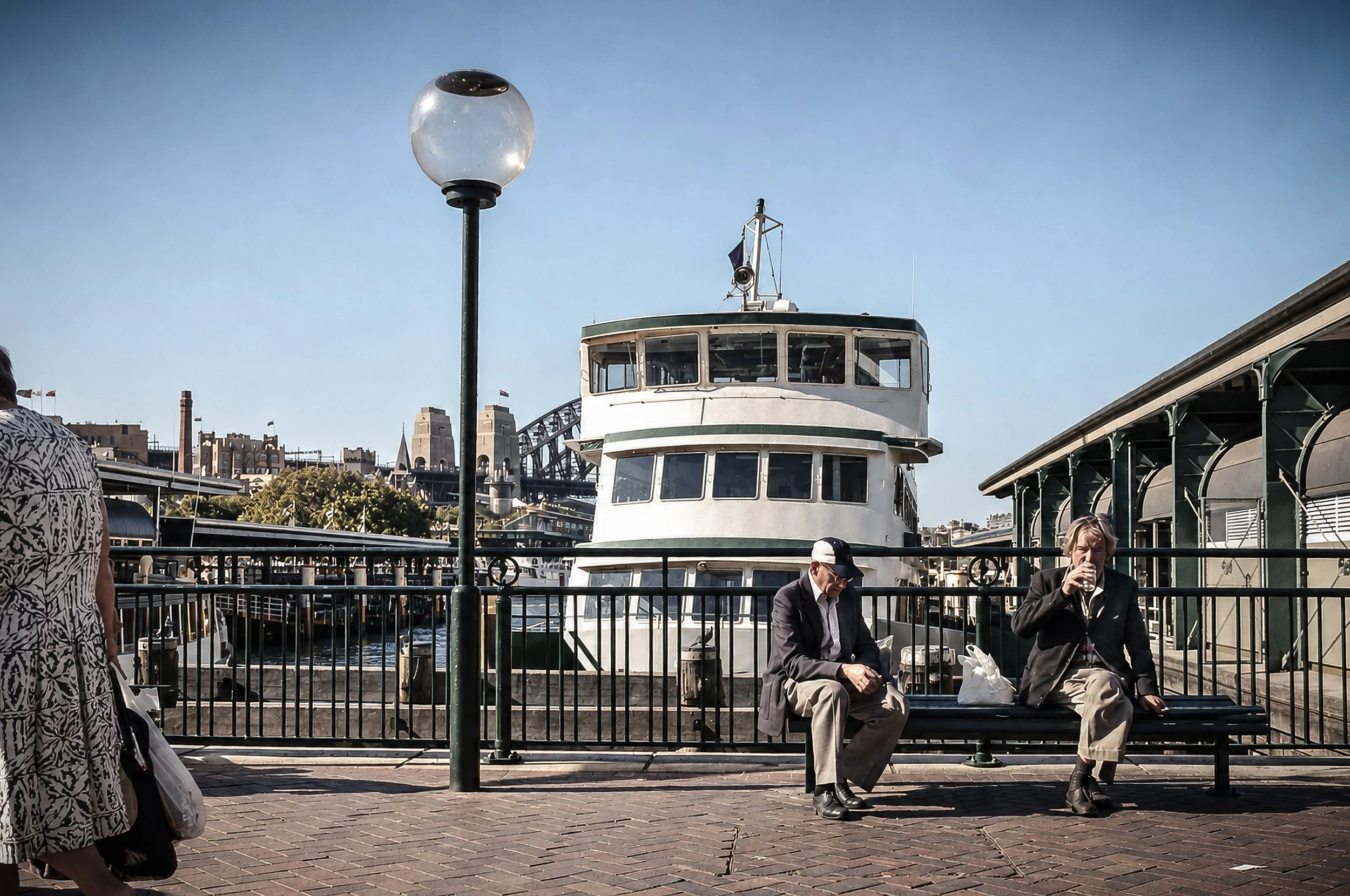 Sydney Harbour Ferry - A ferry approaches the Sydney waterfront as people pause beside the harbour, with the city's architecture and waterfront activity forming a distinctly Sydney backdrop. A quiet observational photograph capturing everyday life be