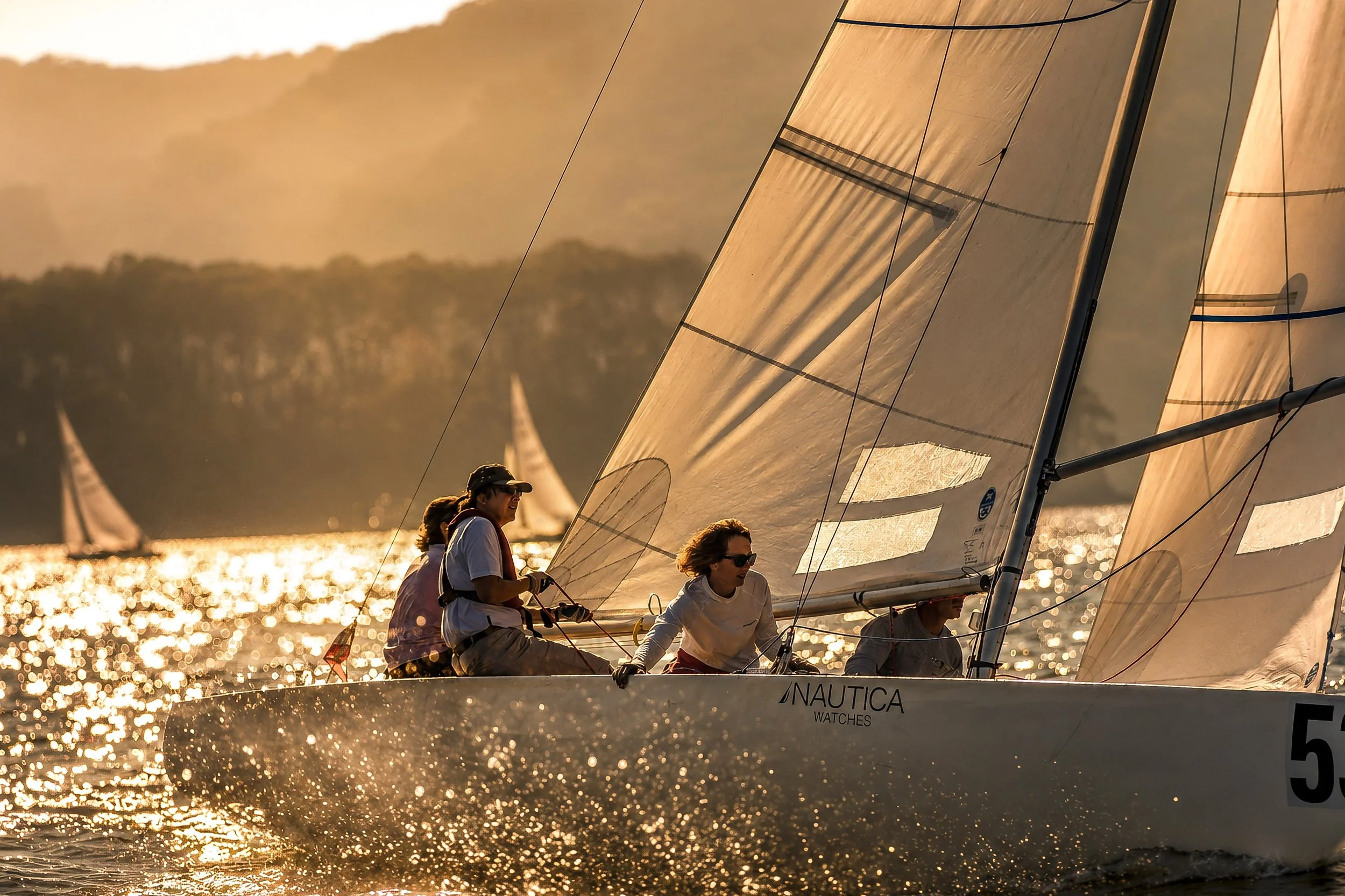 Sailboats on the water during sunset, with multiple sailors adjusting the sails as the sunlight reflects off the water.