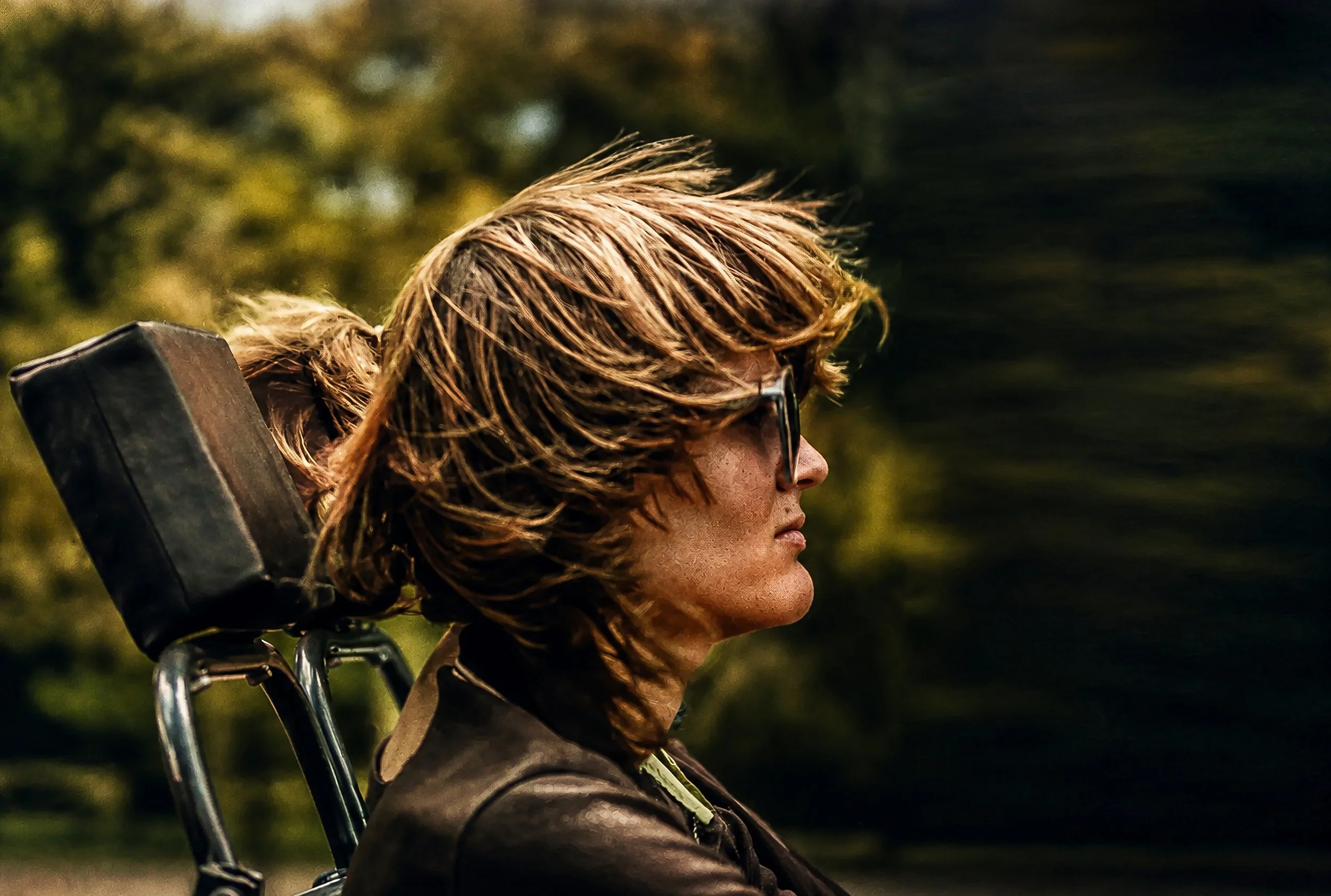 Profile of a woman with short brown hair and sunglasses, carrying a backpack, outdoors with blurred greenery in the background.