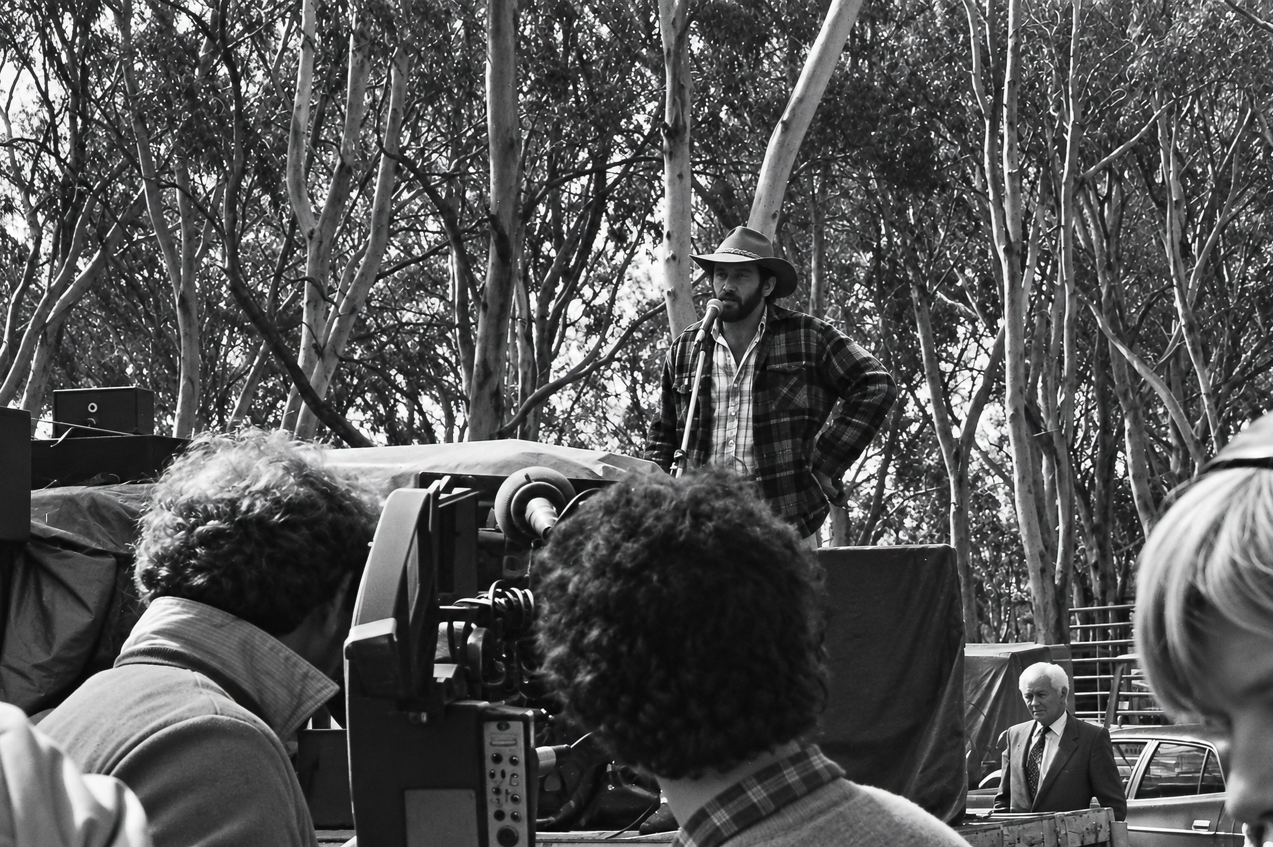 The Speaker - A High Country cattleman addresses the gathering from an improvised platform among the eucalyptus trees. In the foreground, a large television camera records the speech, providing a glimpse of the media presence surrounding the protest.