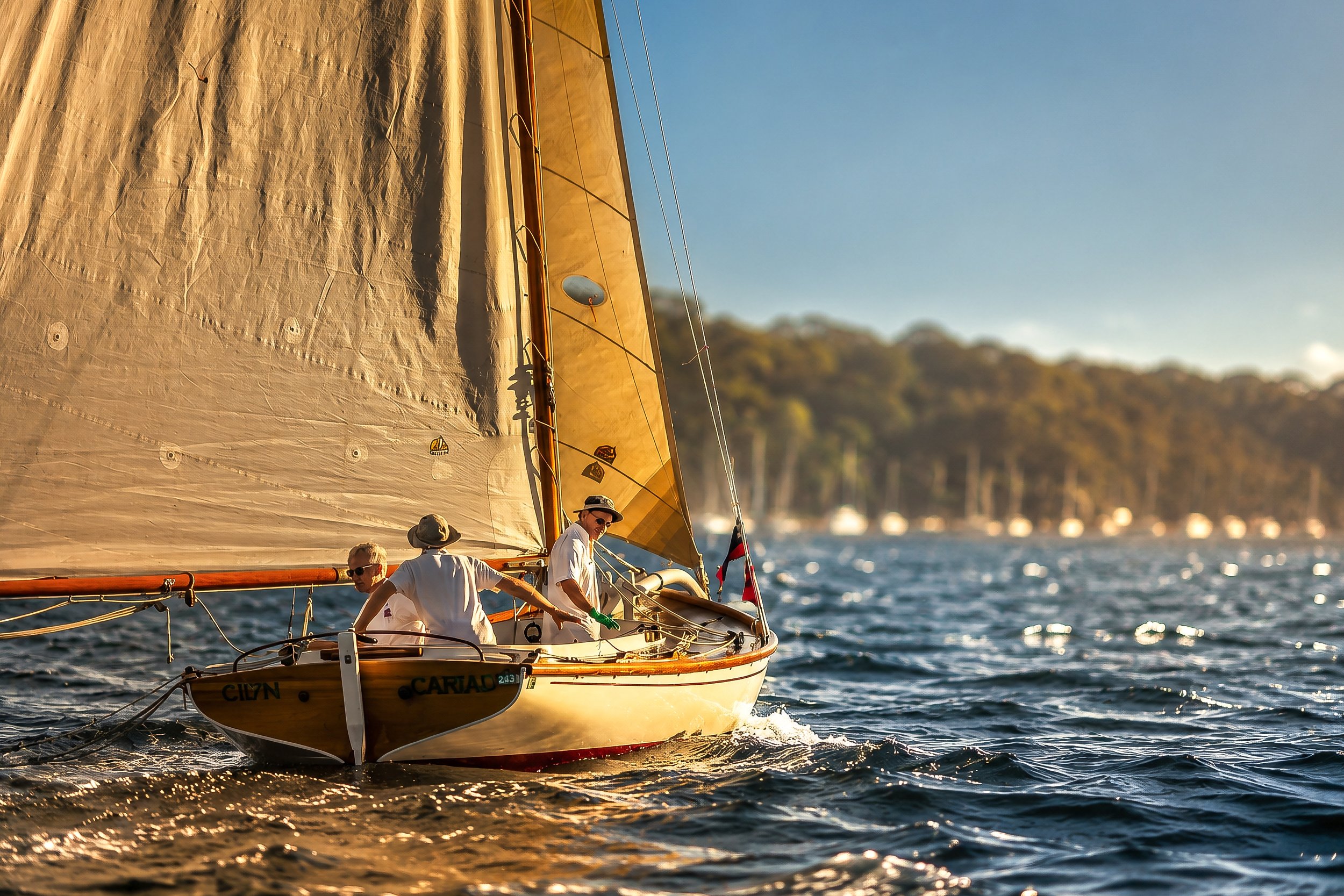 People sailing a boat with a large tan sail on a body of water during sunset, with a forested shoreline in the background.