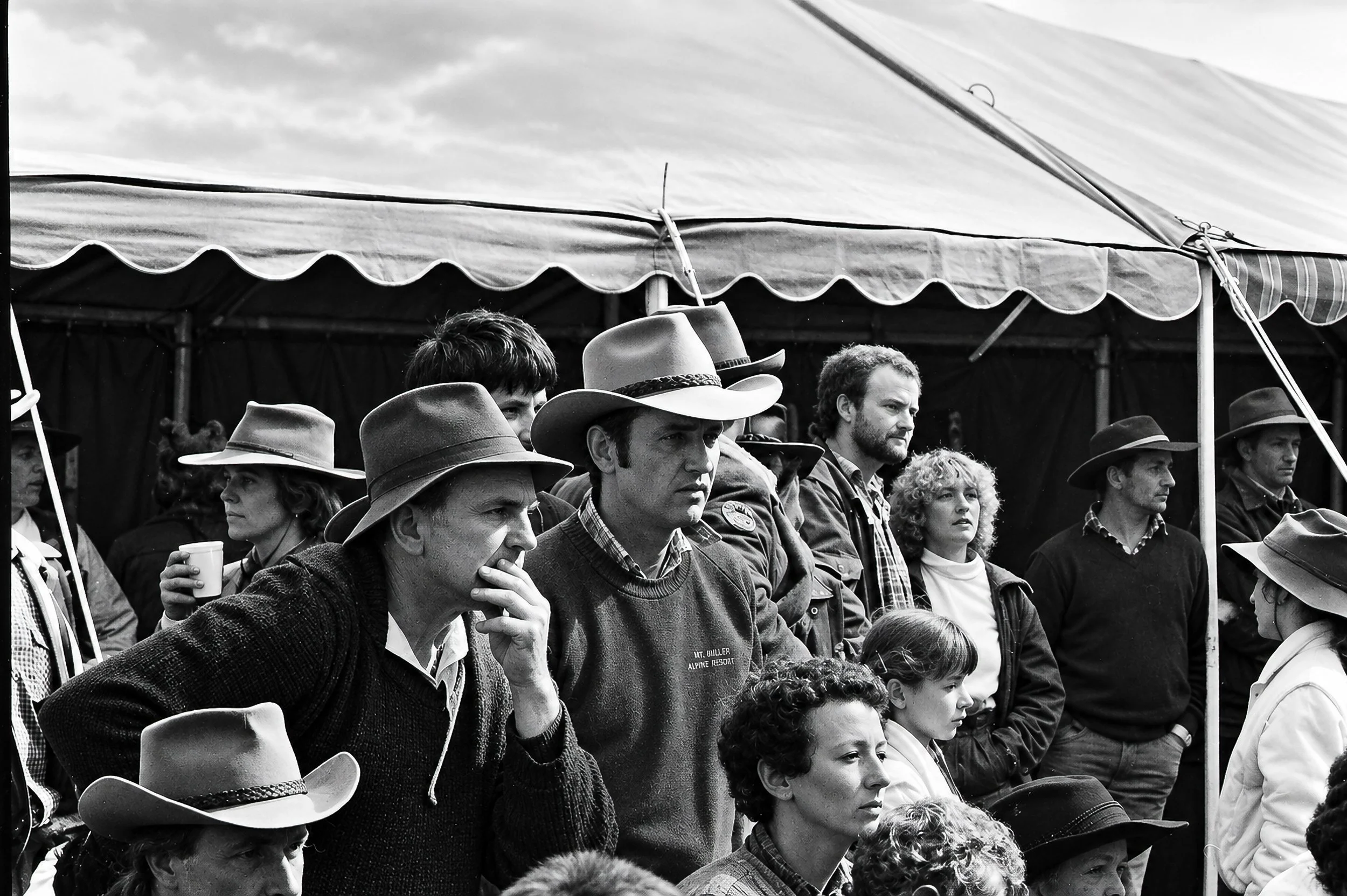 Waiting Beneath the Tent - Cattlemen, families and supporters gather beneath a large canvas tent. Hats, faces and expressions emerge from the deep shade as people wait for the day's events to unfold.