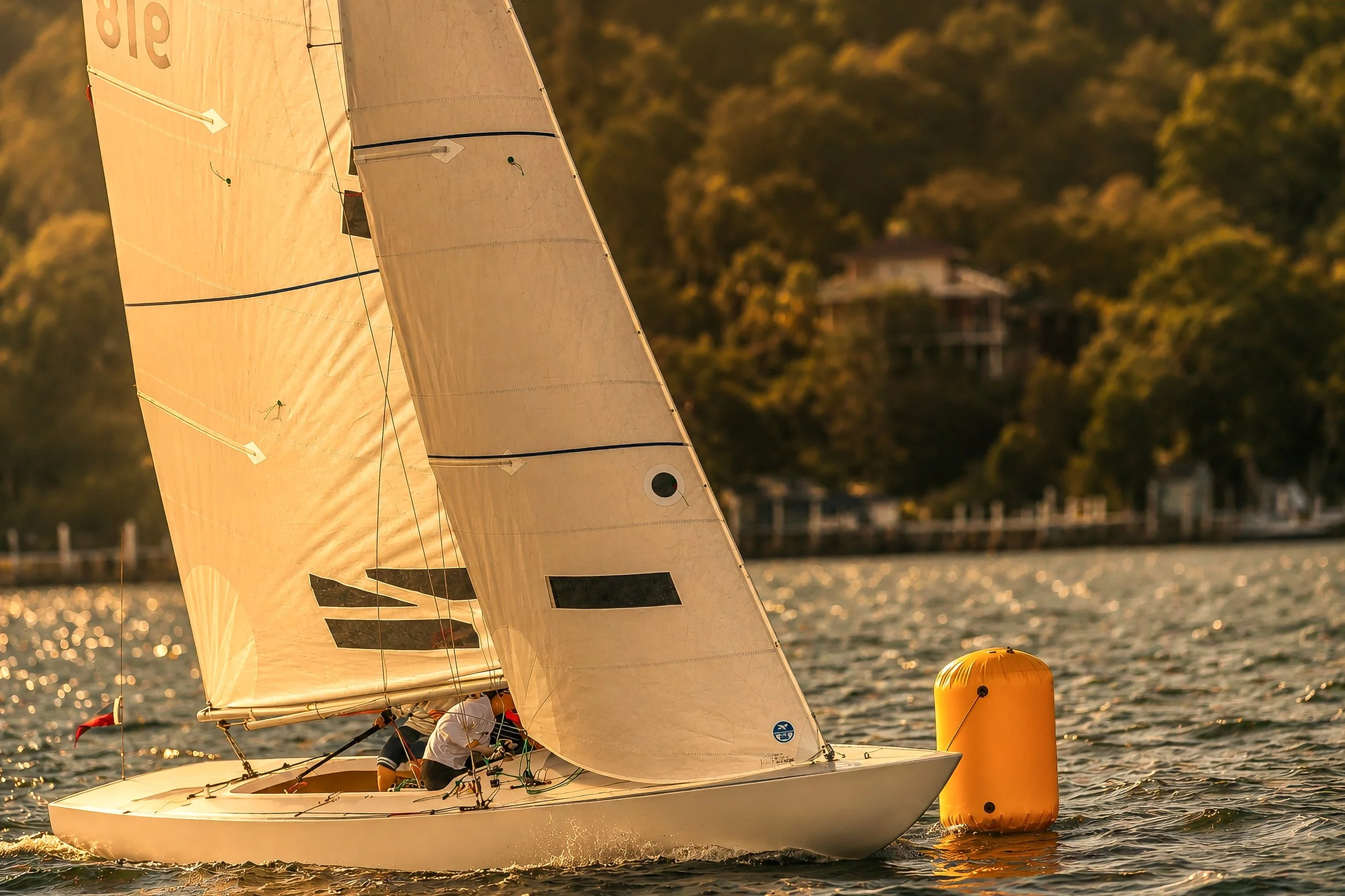 A sailboat racing on the water with two people onboard, near a large orange buoy during sunset, with a house among trees in the background.