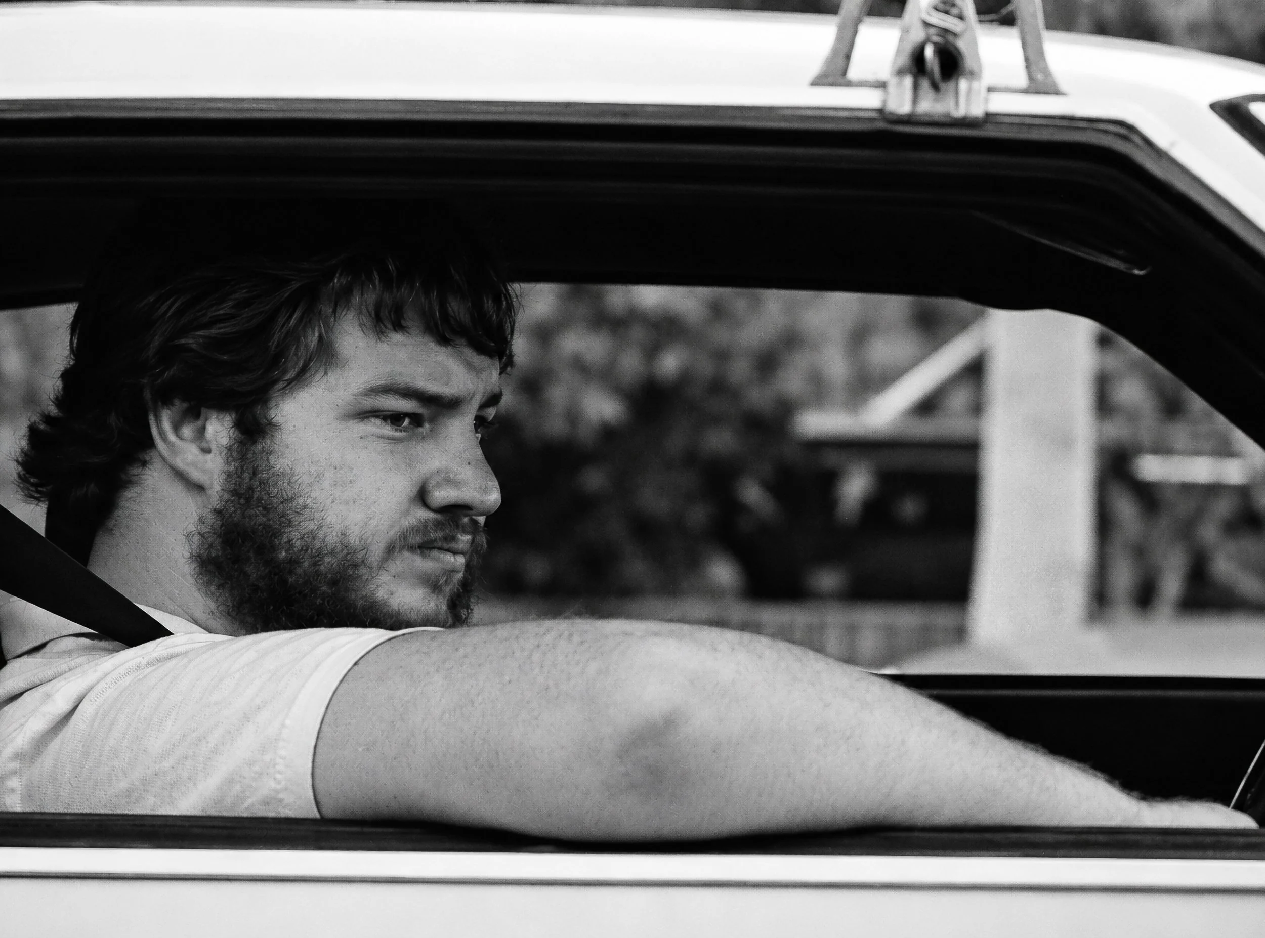 The Young Driver - A young bearded driver rests his arm along the window as he looks towards the road. The simple composition of face, arm, car interior and open window captures the relaxed character of an ordinary Sydney traffic moment.