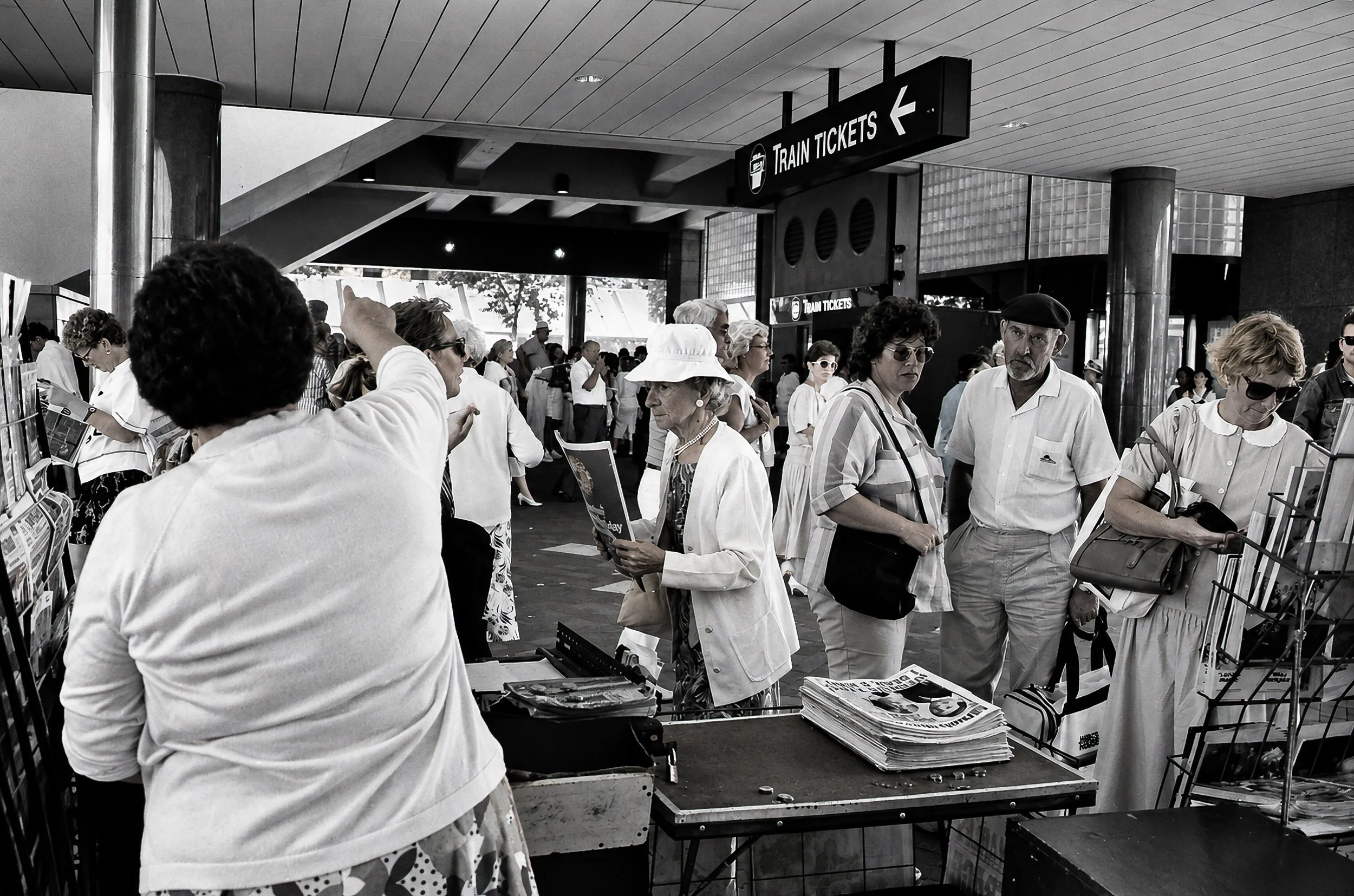 The Waiting Crowd - A busy station concourse filled with passengers, newspapers, luggage and movement. The Train Tickets sign and the period clothing and advertising turn an everyday railway scene into a detailed record of Sydney in the 1980s.
