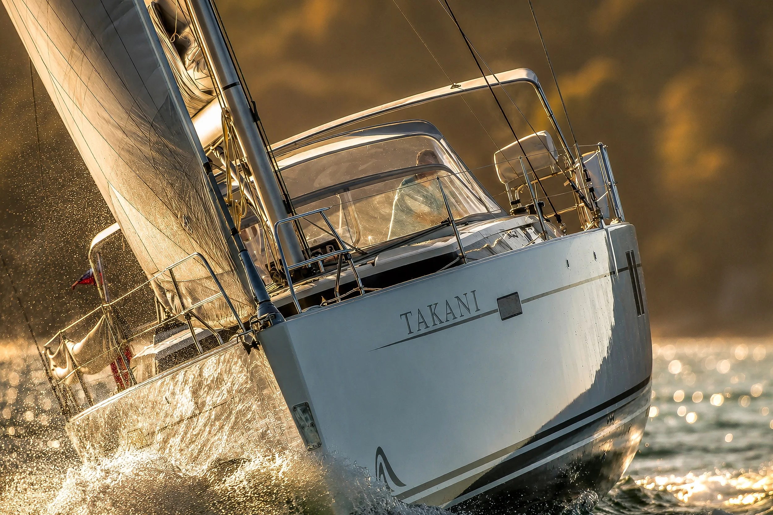 A sailboat with the name TAKANI sailing on water at sunset, with a person visible inside the boat.