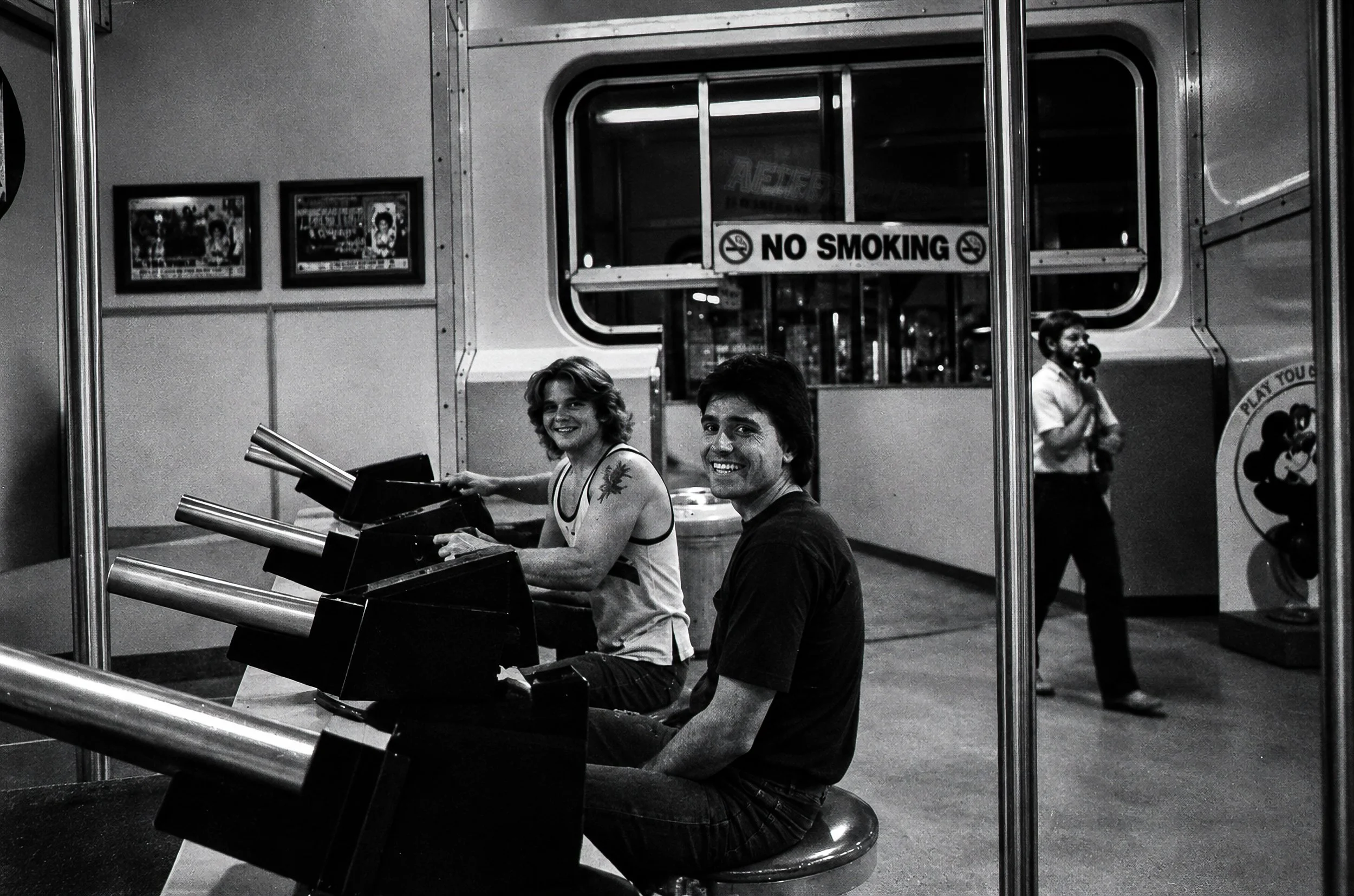 After Hours - Two passengers pause at a shooting-game arcade while another person talks on the telephone in the background. The NO SMOKING sign, machinery and interior details preserve a particularly strong sense of the period.