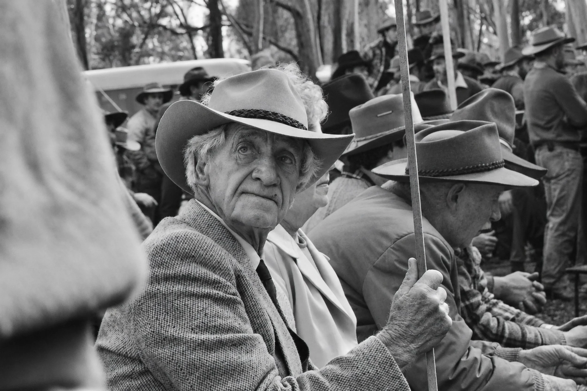 A Face in the Crowd - An older cattleman looks directly towards the camera while holding a protest sign pole. His weathered face, broad-brimmed hat and country clothing provide an intimate portrait within the much larger gathering.