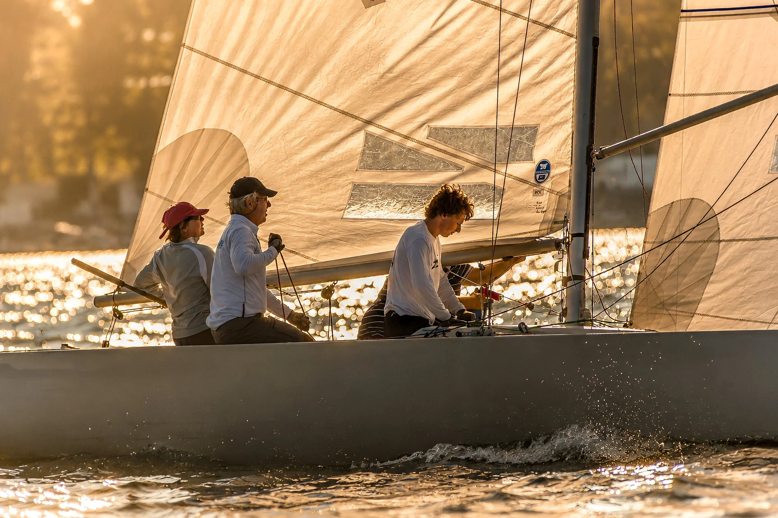 A sailing boat with four people onboard, sailing on water during sunset; the boat's sails are illuminated by the sunlight and the water glows with reflections.