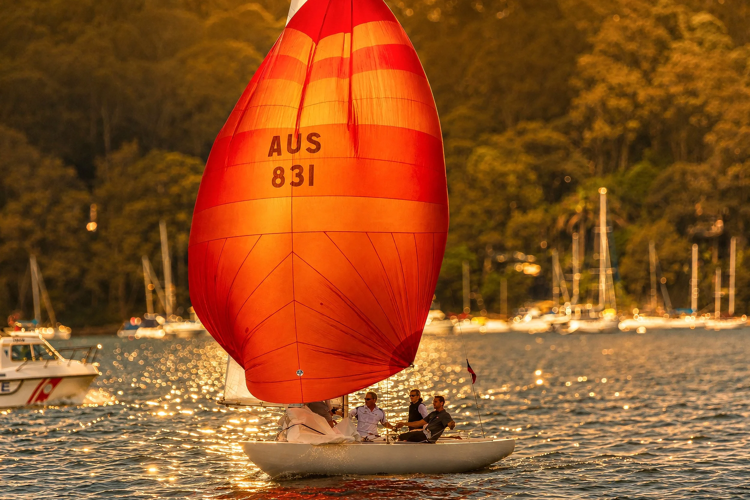 A sailboat with a large red and orange striped spinnaker sail on a body of water during sunset, with reflections on the water and a background of trees and sailboats.