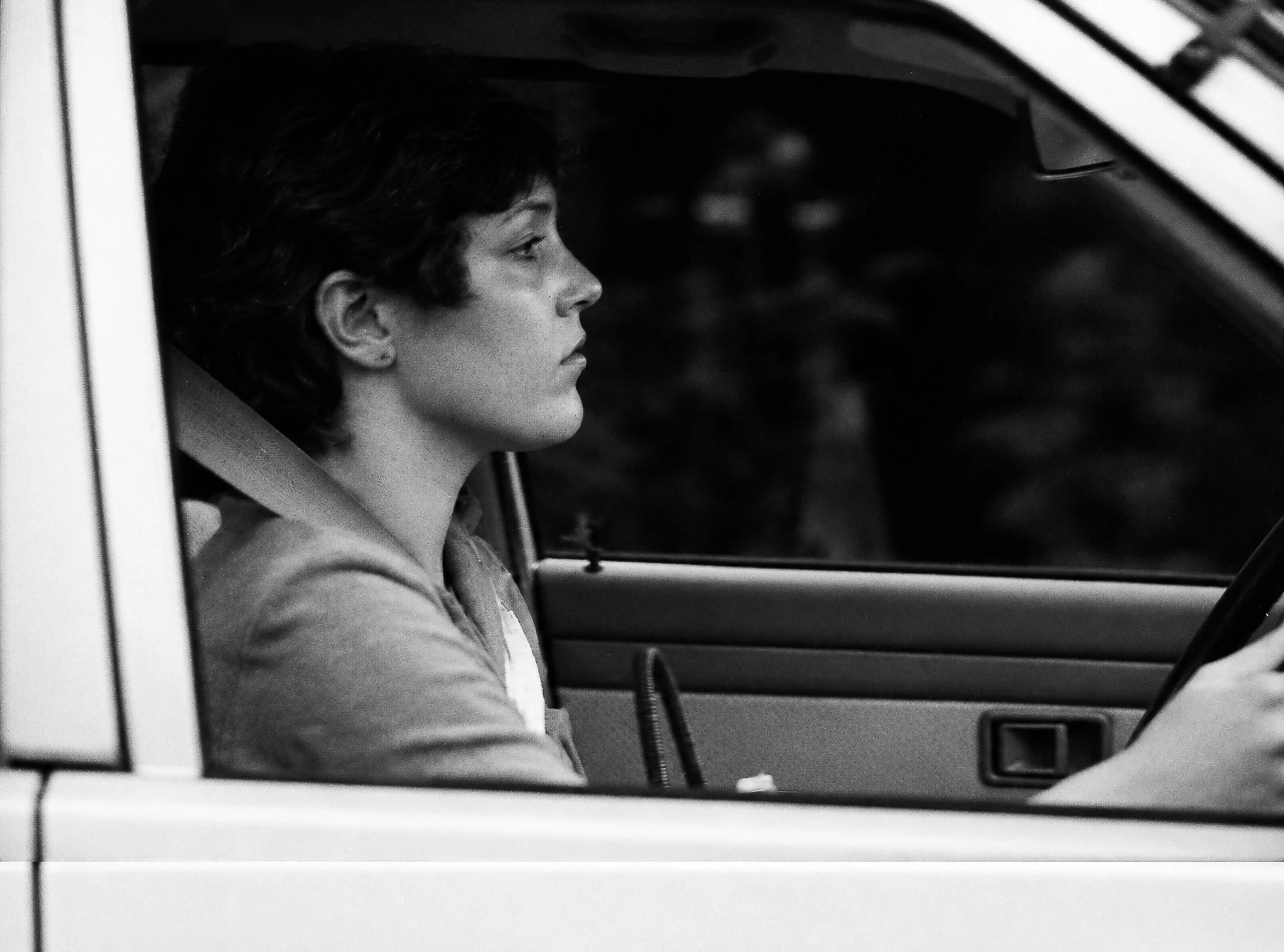 Quietly Passing - A woman driver sits in profile, concentrating on the road ahead. With its dark interior, restrained expression and simple composition, the photograph provides a quieter moment within the collection.