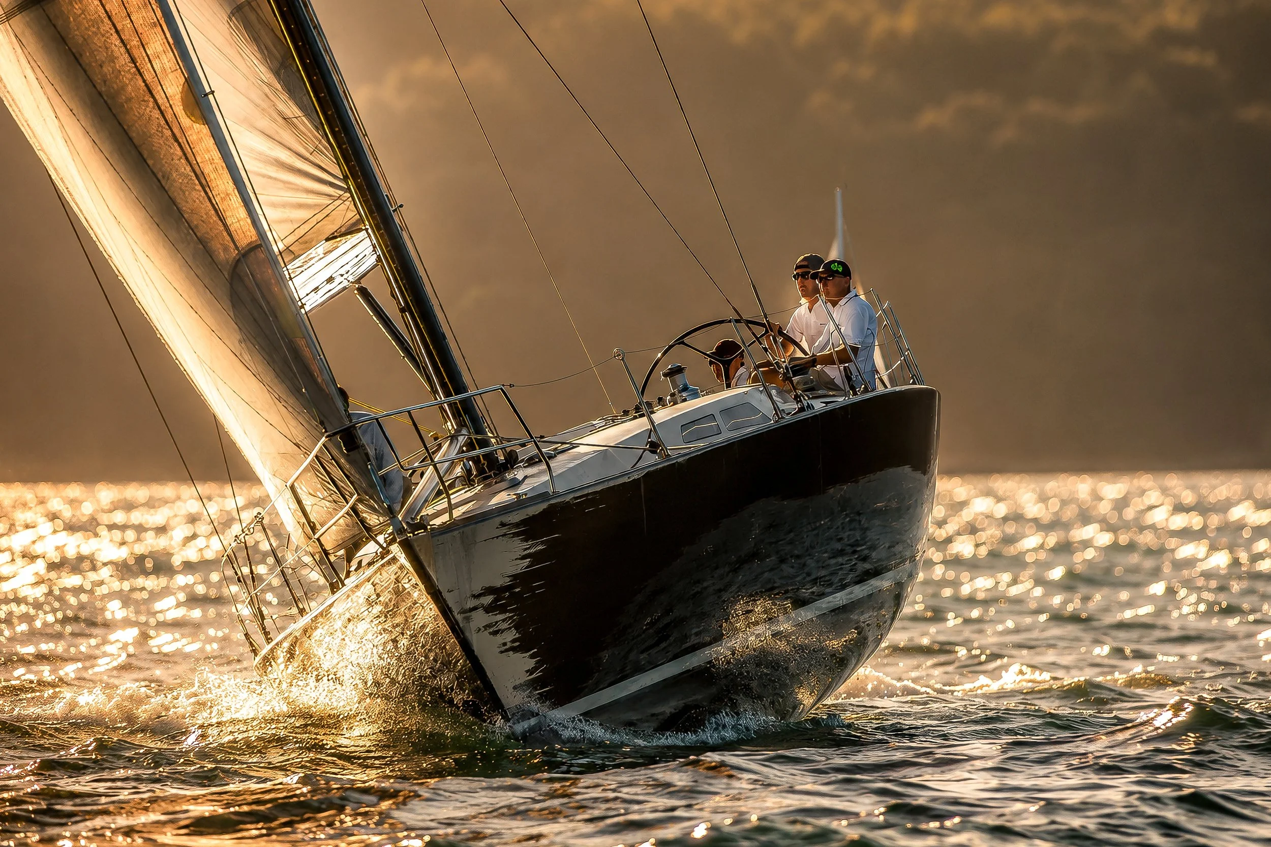 A sailboat moving through water at sunset with crew members onboard, the reflection of the sun shimmering on the water.