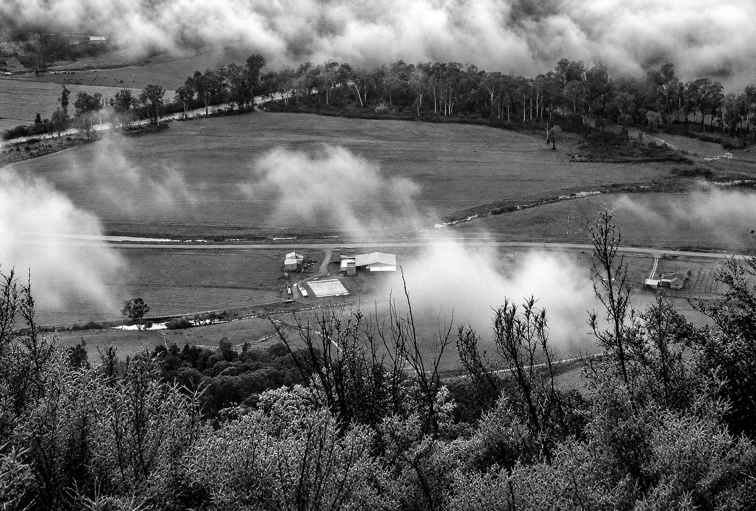 The Valley Below - Mist drifts across open farmland and around a small rural settlement below the mountain viewpoint. Roads, fields, trees and buildings appear briefly through the cloud, providing a glimpse of the human landscape beneath the weather.