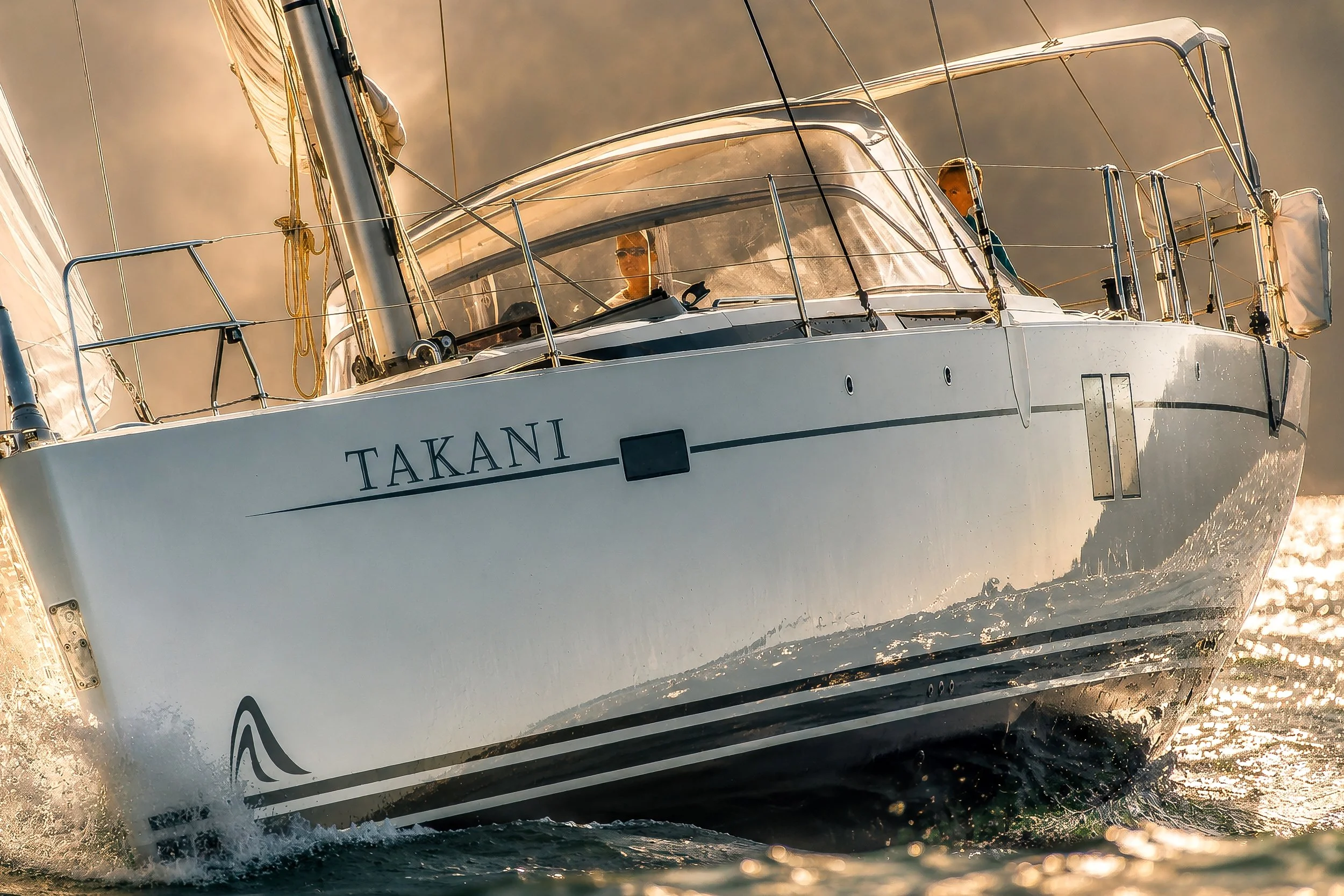 A sailboat named TAKANI sailing on water with its sails up, with two people visible inside the cabin, and the photo taken from the water level.
