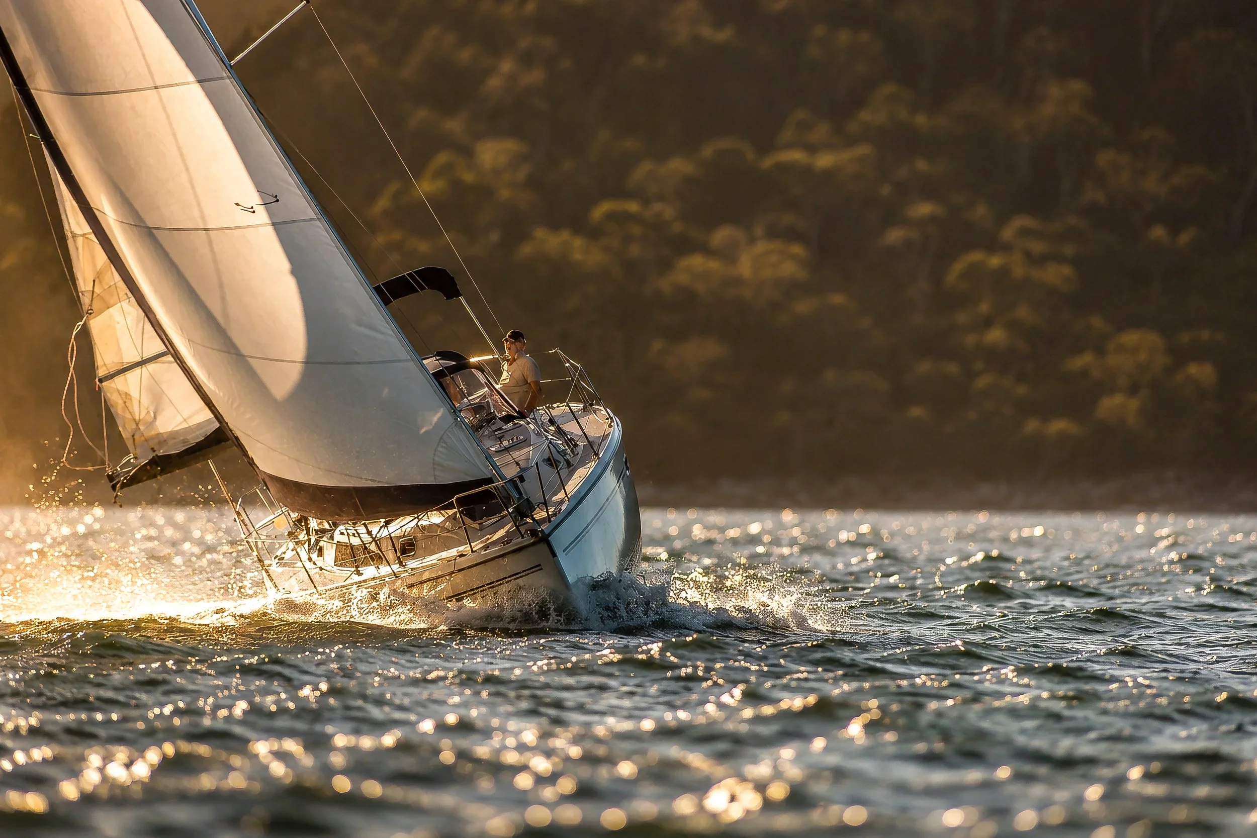 A sailboat moving quickly across a body of water during sunset, with a person on board, surrounded by sparkling reflections on the water surface and a blurred tree-lined shoreline in the background.