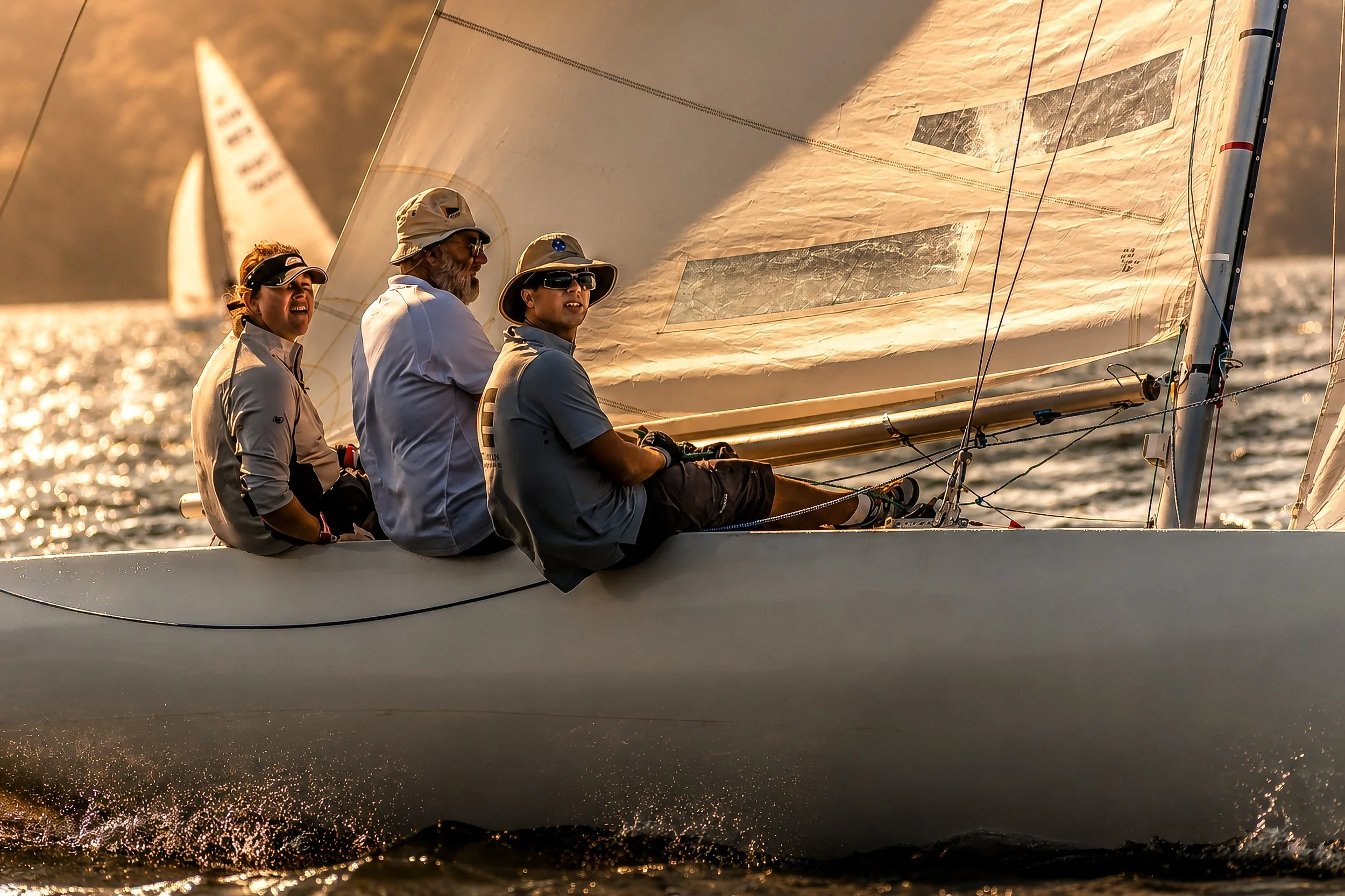 Four people sailing on a boat in the water during sunset, with sails raised and focused on steering.