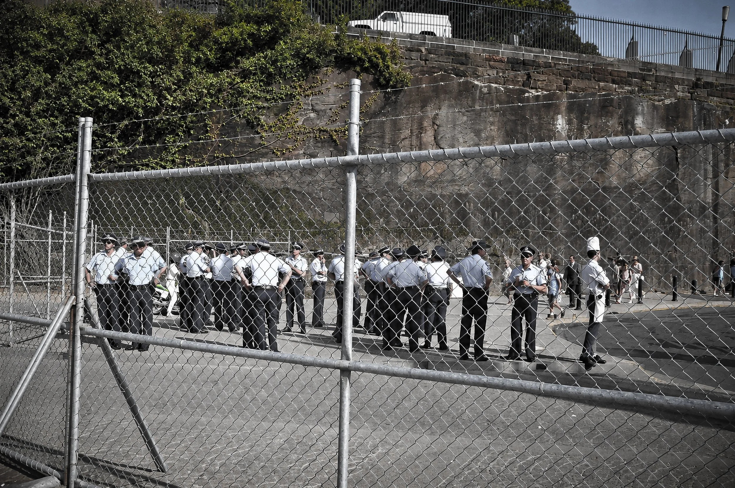 Police and Crowd Behind the Fence - A group of police officers and members of the public gather behind a chain-link fence. The strong geometric pattern of the fence contrasts with the people beyond it, creating a compelling documentary photograph of 