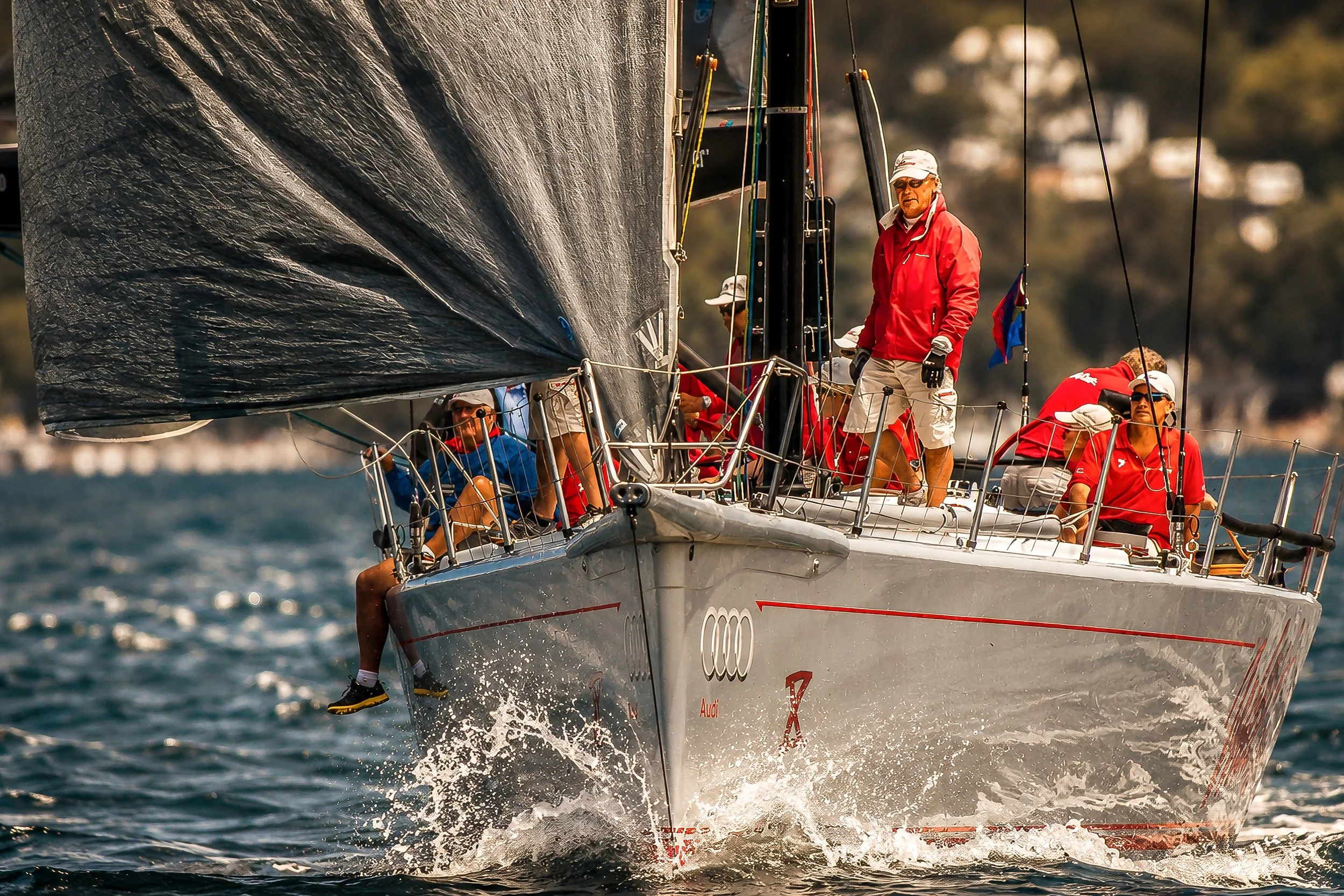 A sailing yacht with a group of people on board, navigating through the water with the sail caught in the wind. The crew members are wearing life jackets and hats, and the yacht is moving at a high speed, creating a splash in the water.