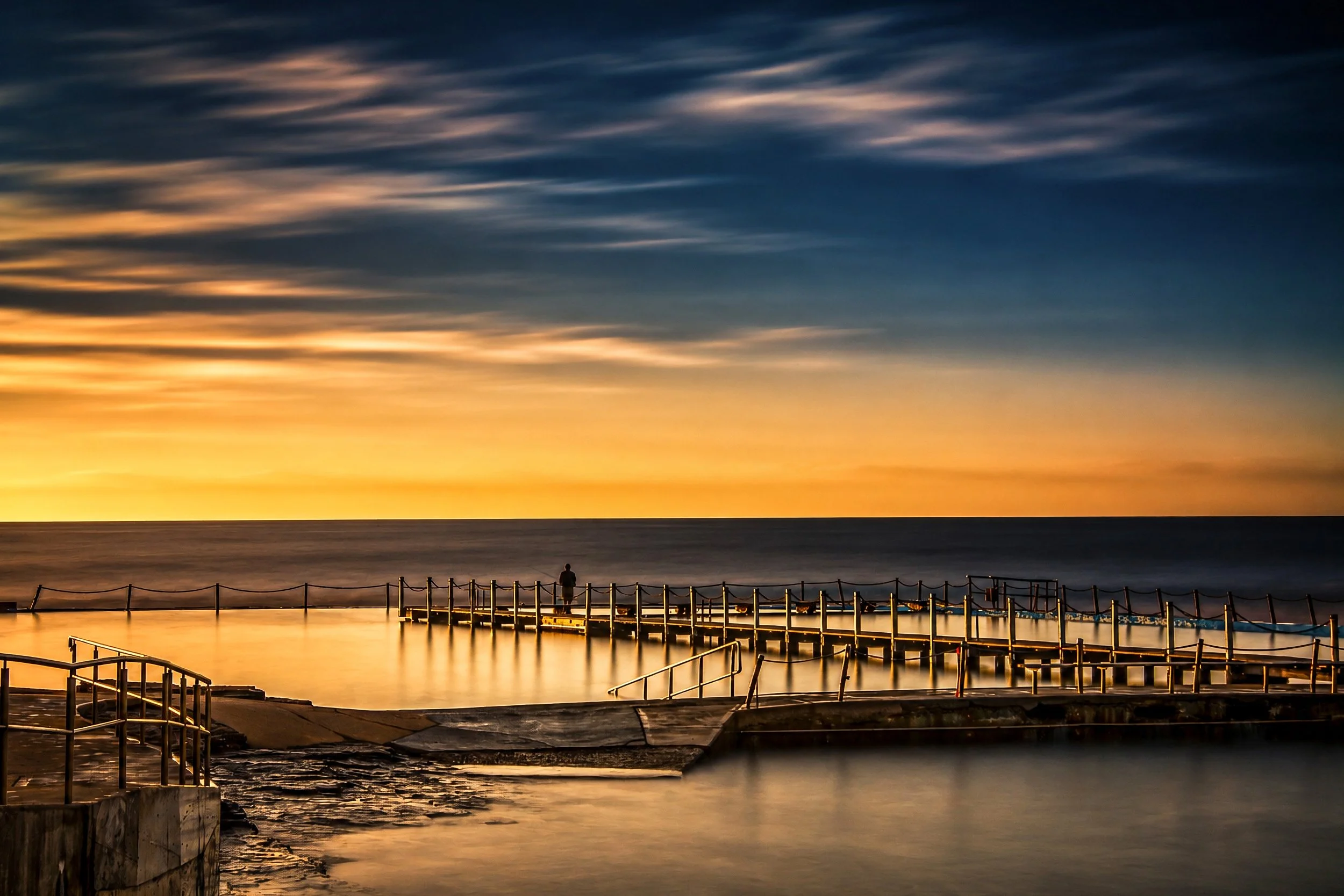 A tranquil ocean pool and timber jetty glow beneath the first light of dawn.

