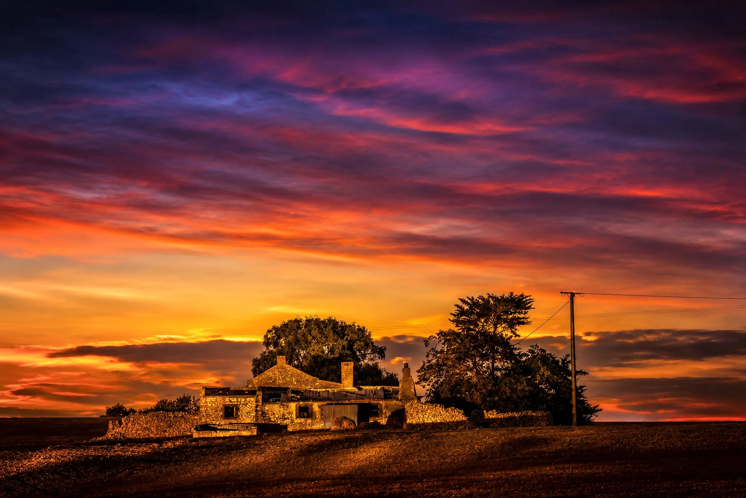 An old rural farmhouse silhouetted beneath a spectacular crimson sunset sky.
