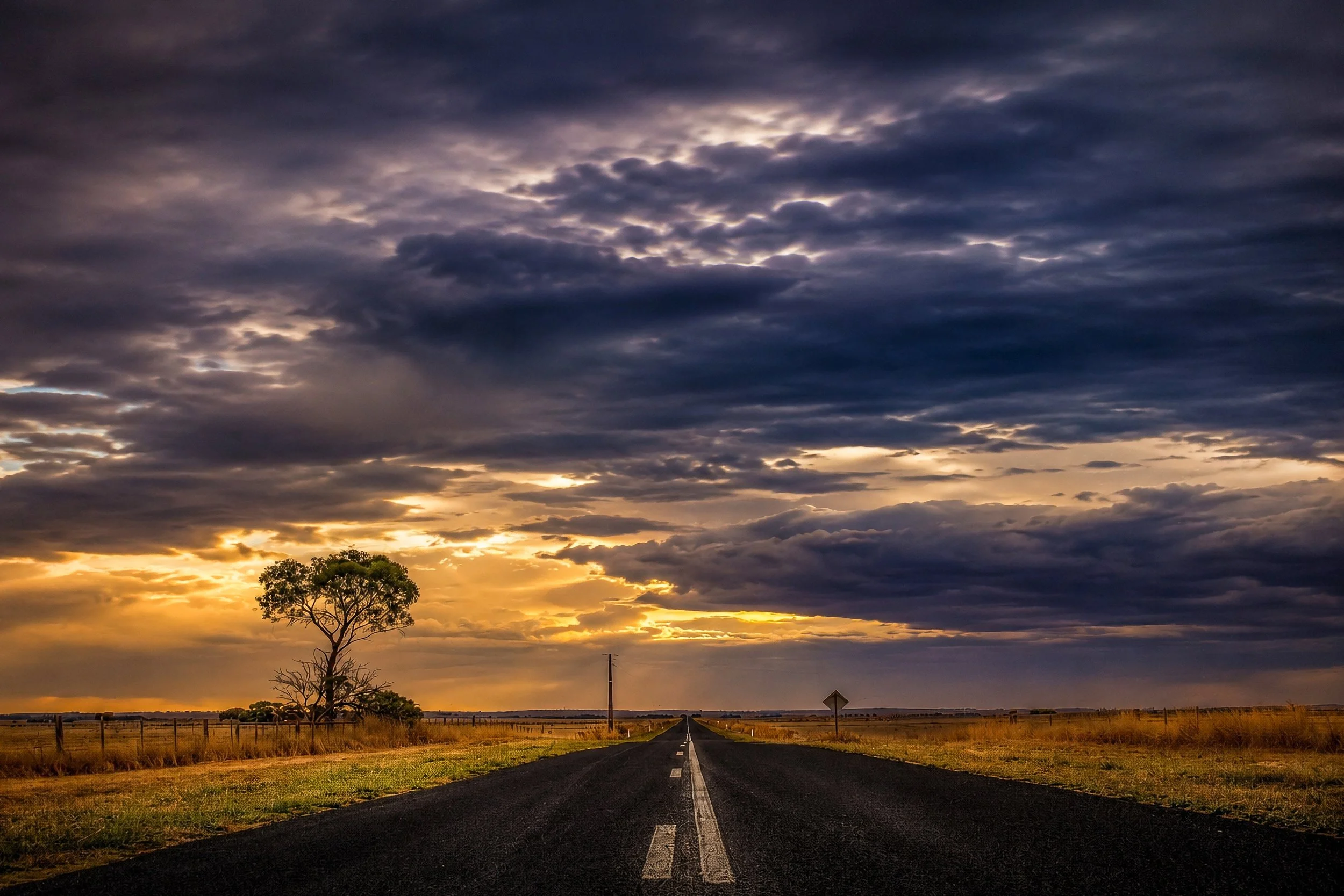 A lonely country road disappears beneath dramatic storm clouds across the Australian plains.
