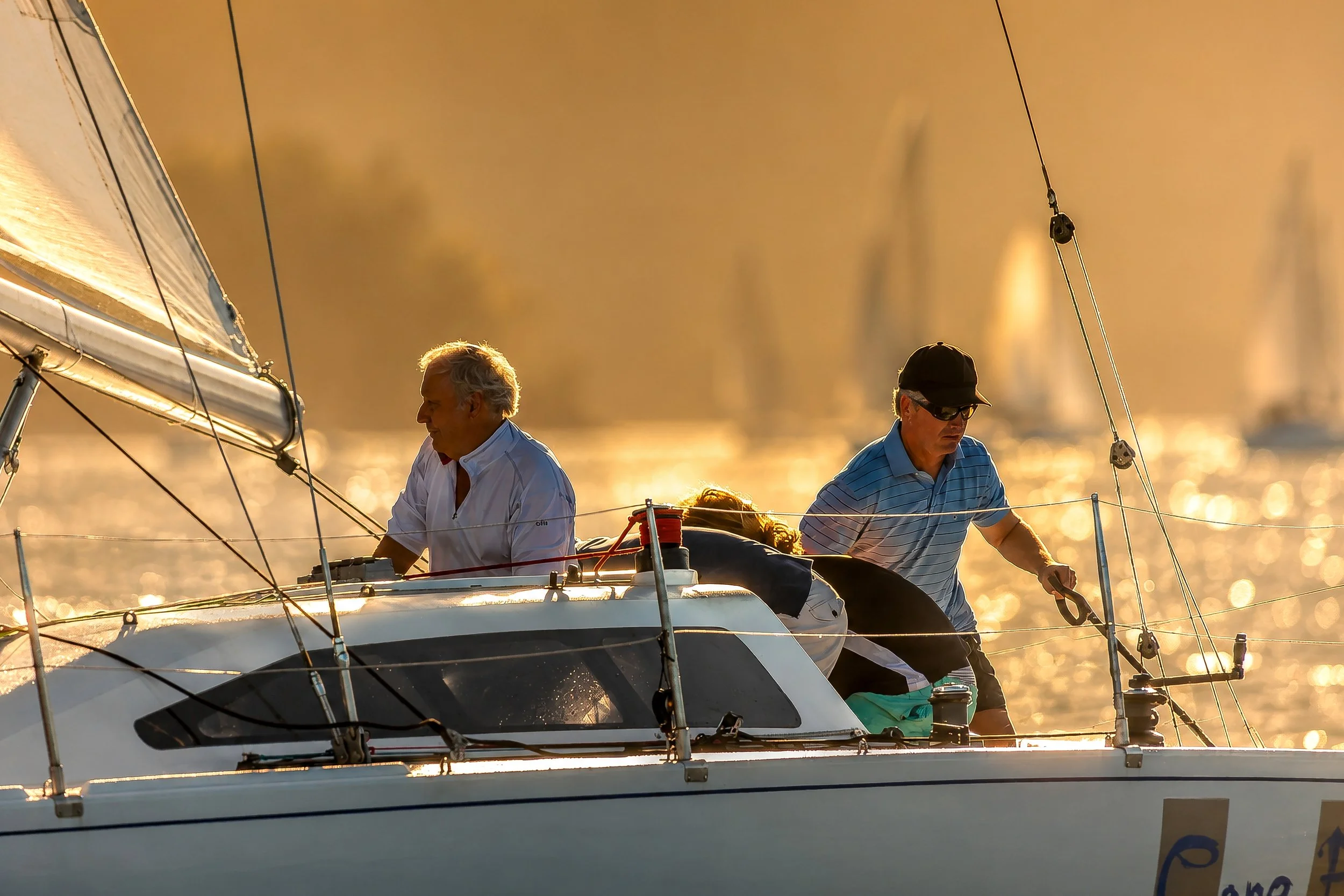 Two men sailing a boat during sunset, with the sail visible and the water reflecting the sunlight.