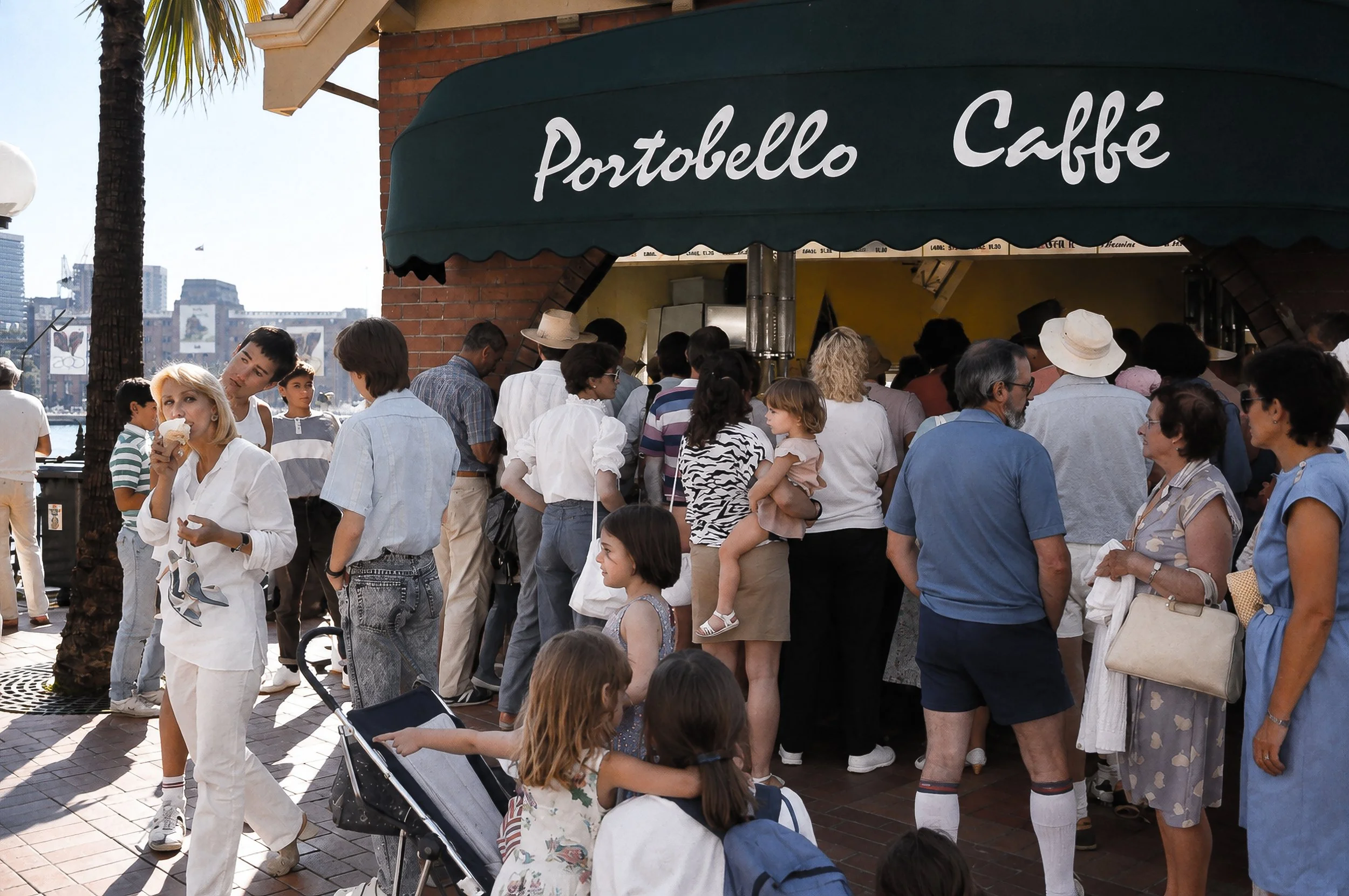 Portobello Caffé - Sydney Street Life - A wider view of the crowd outside Portobello Caffé reveals the café as a meeting place within the busy waterfront environment. People, clothing, signage, architecture and the harbour combine to create a detaile