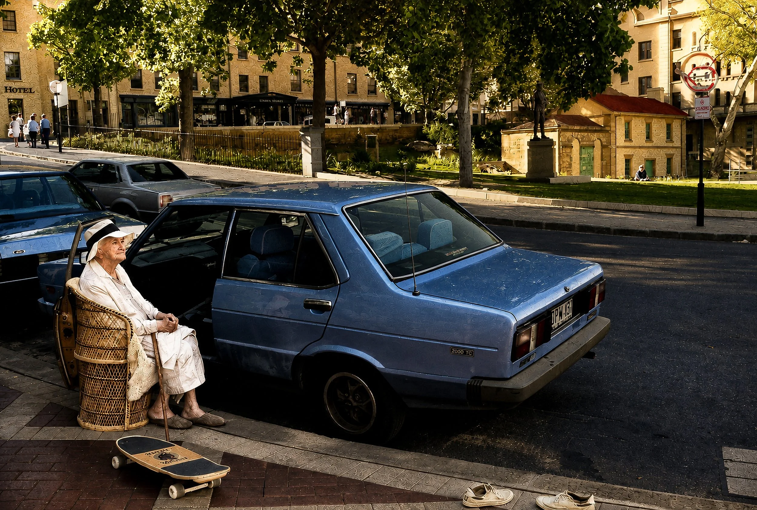 An elderly woman sitting on a chair near parked cars on a city street, with a skateboard on the ground nearby, trees and buildings in the background.