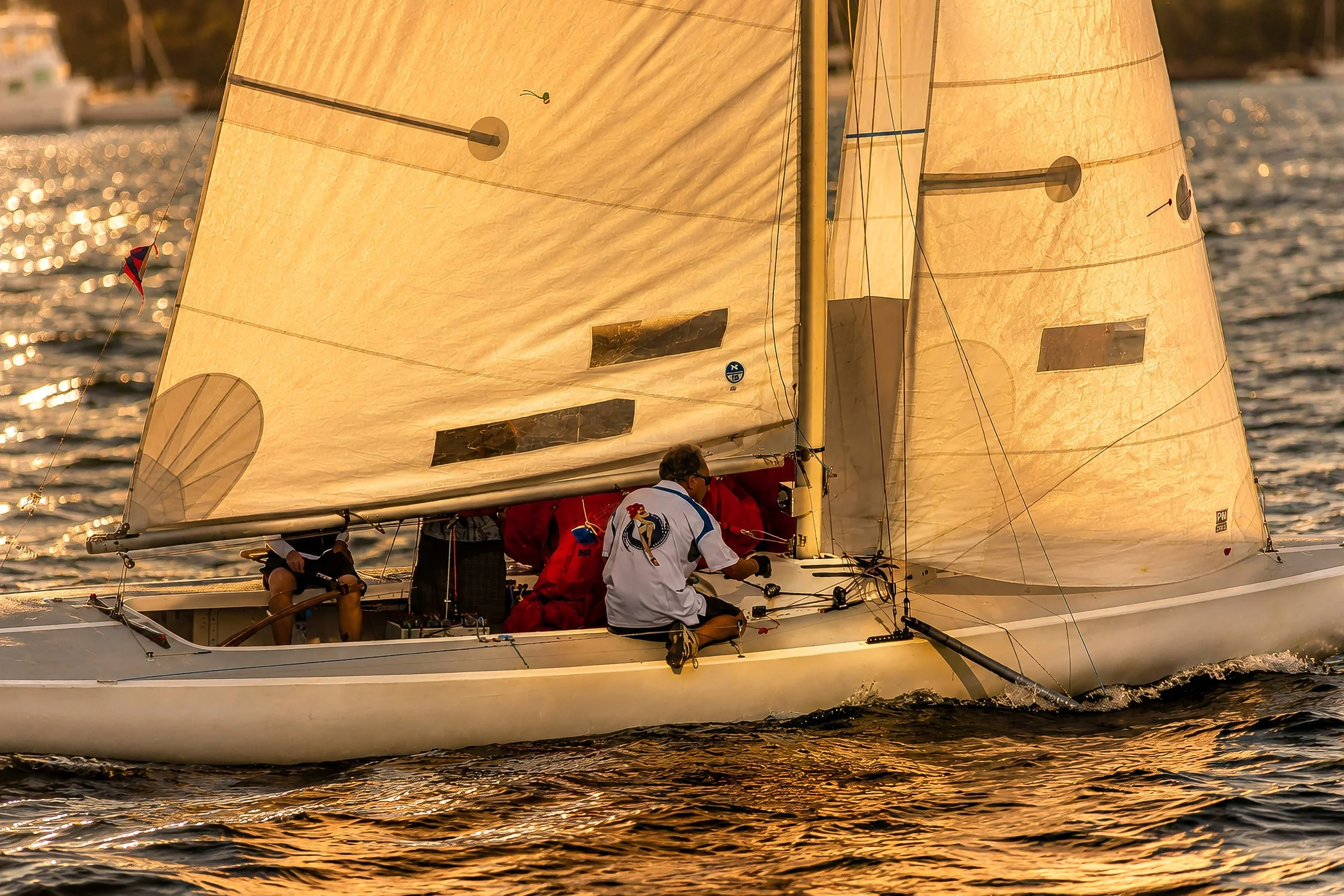 A sailboat with a golden-colored sail sailing on water during sunset, with three people aboard.