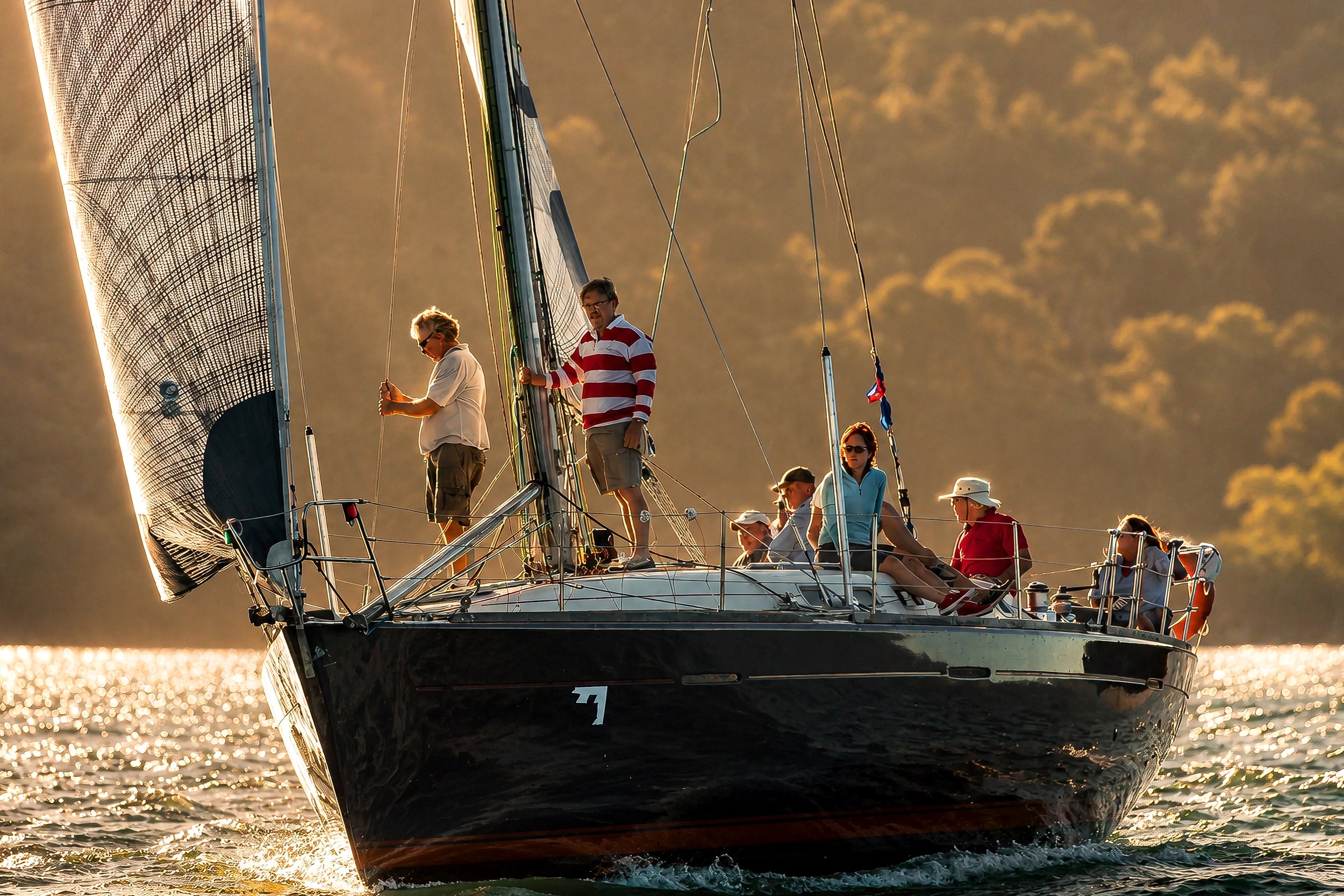 A sailing yacht with multiple people on board sailing during sunset, with the sun illuminating the sails and creating reflections on the water.