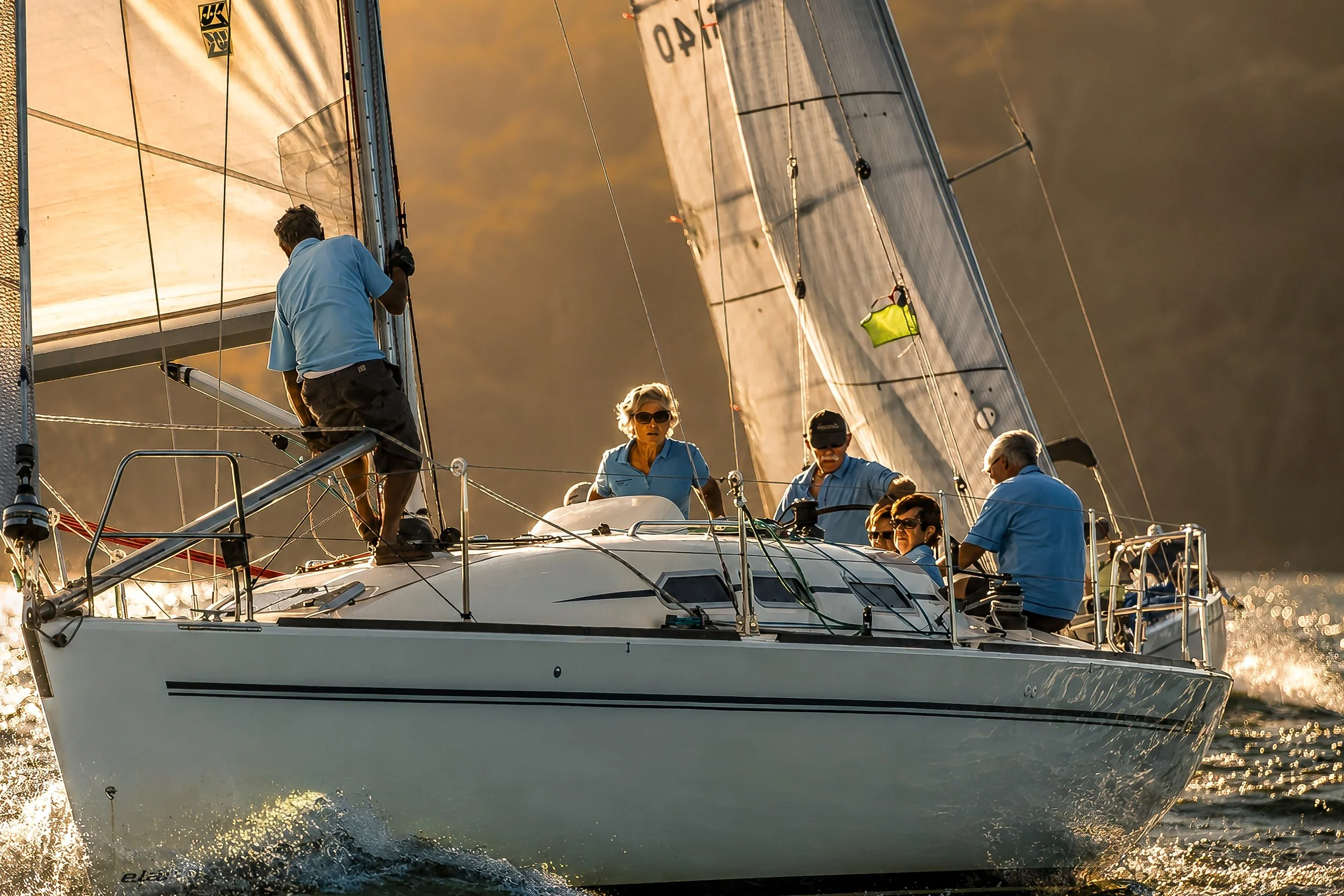 A sailboat with a crew of six people, four men and two women, sailing on calm waters during sunset or sunrise, with the sails catching the wind and the crew adjusting the ropes.
