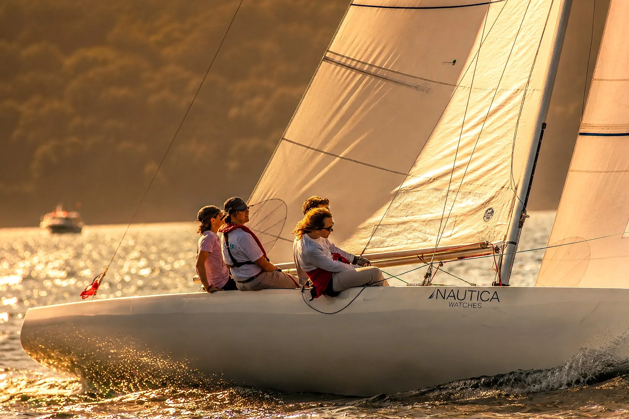 A group of four women sailing on a boat, with sunlight reflecting on the water and a boat in the background.