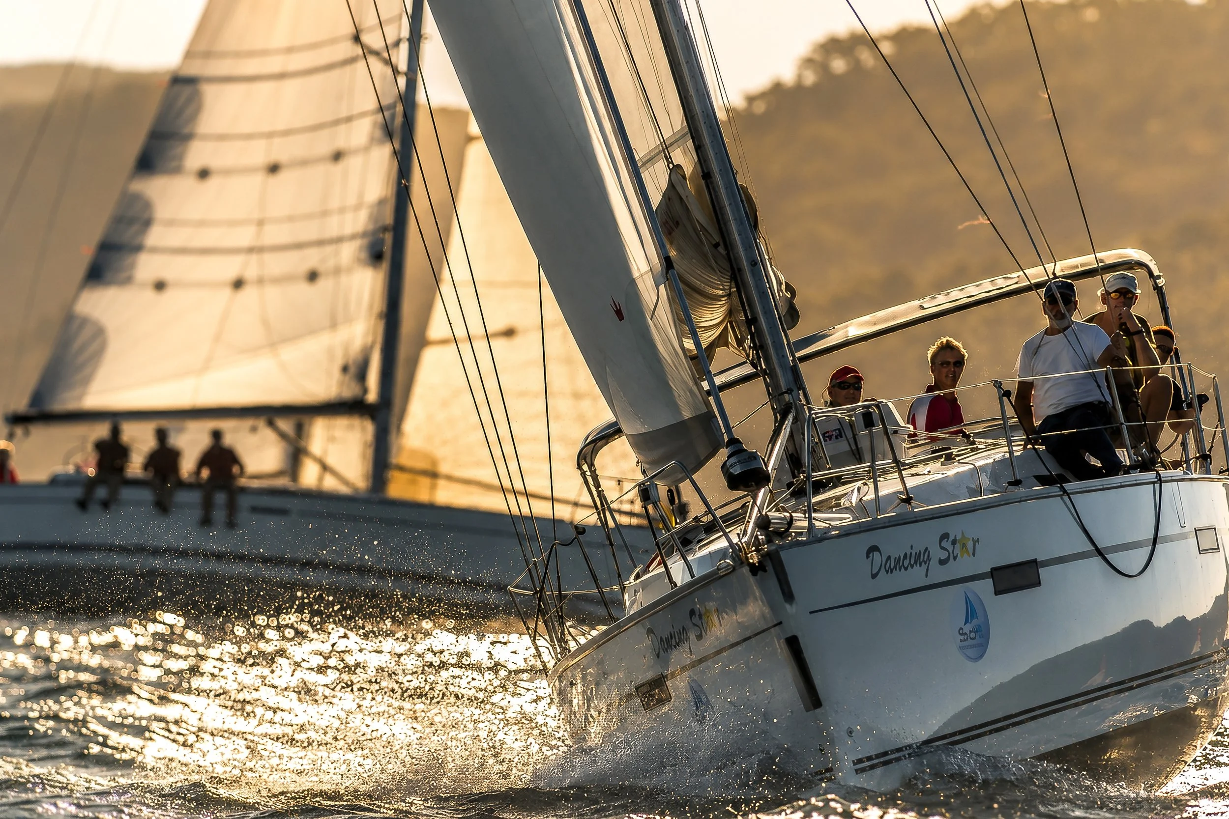 A sailboat named Dancing Star sailing on water at sunset with crew members on deck, sunlight reflecting off the water, and sails unfurled.