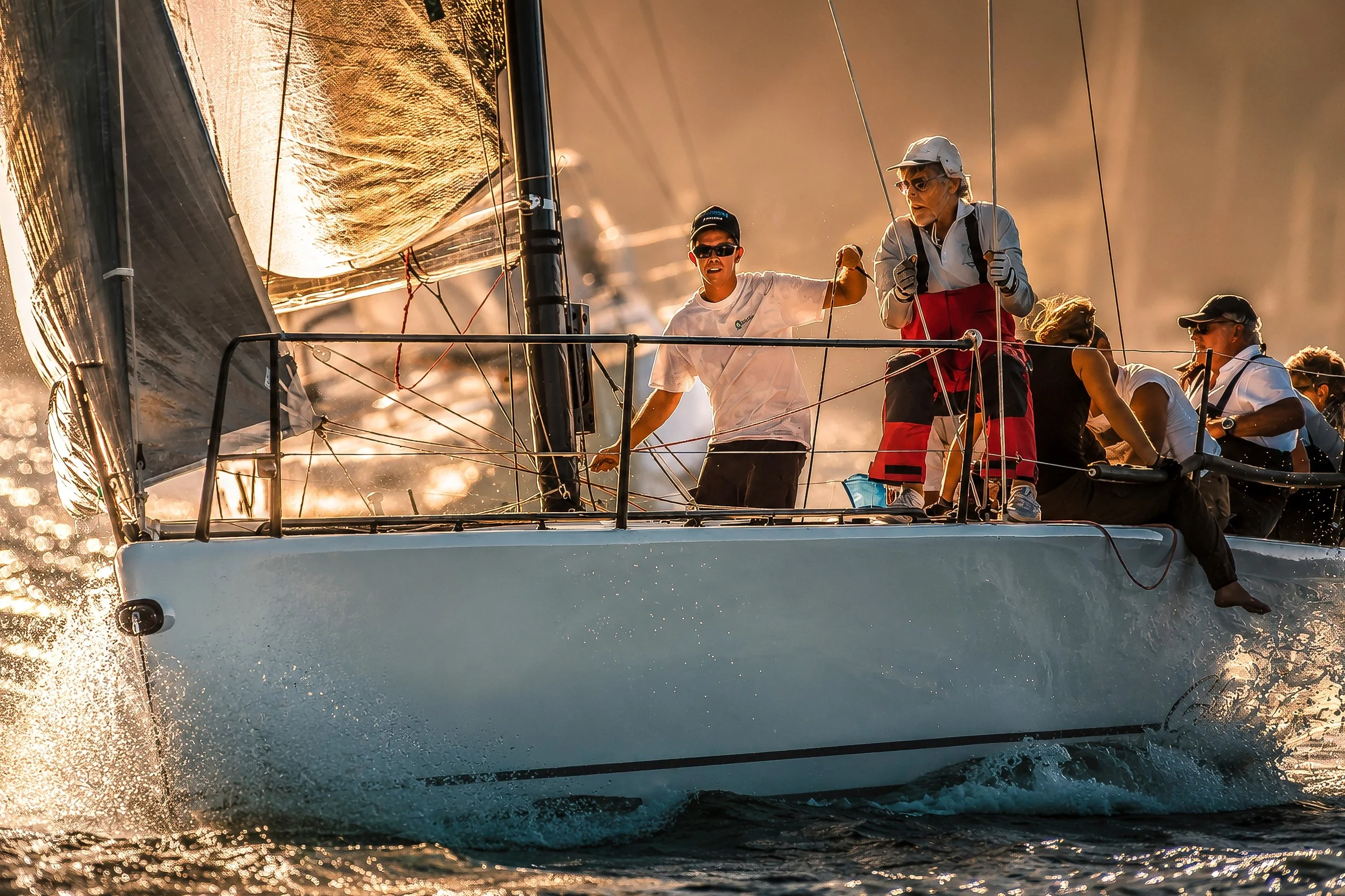 A group of people sailing on a boat during sunset, with calm waters and reflective sunlight on the sails.