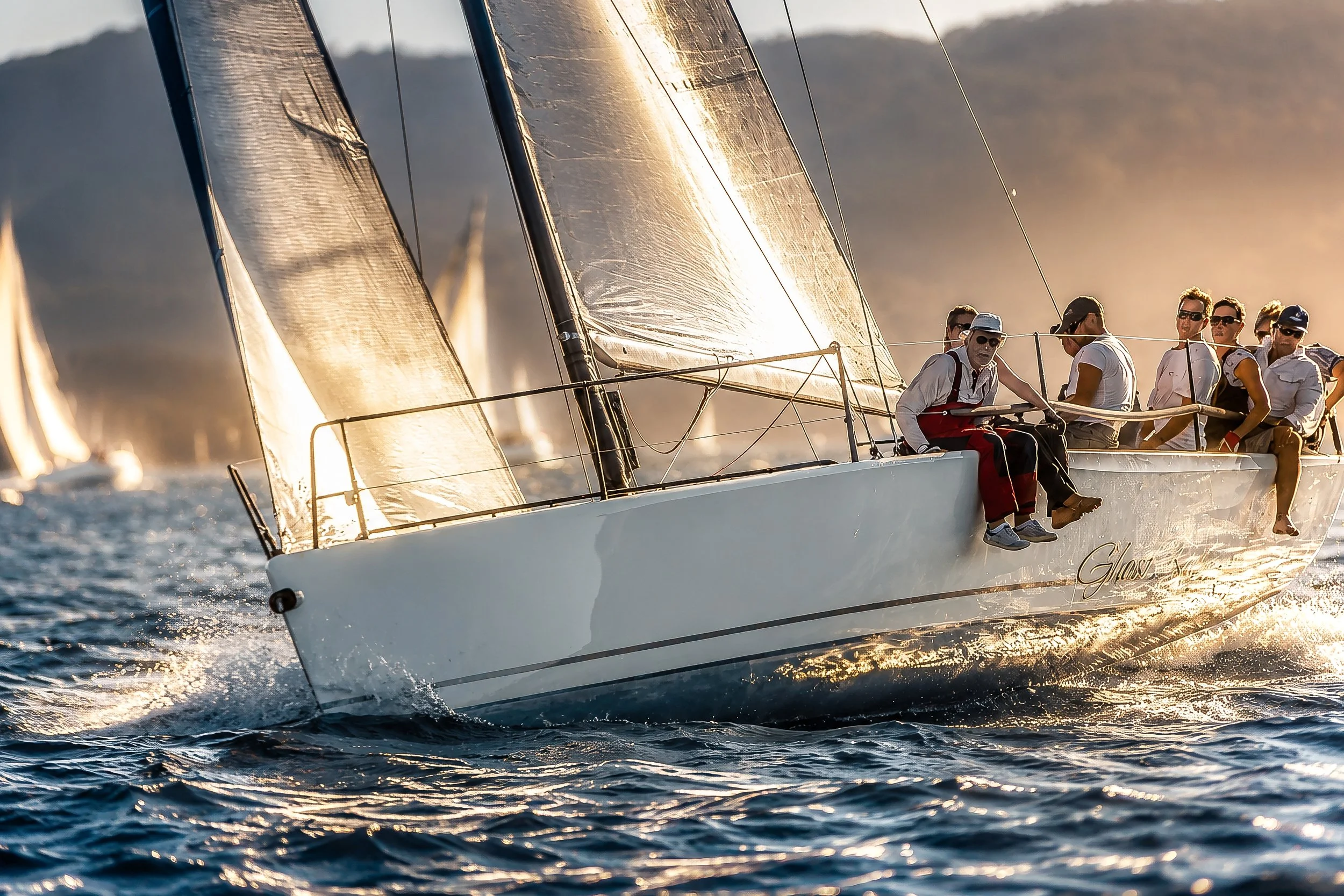A sailboat with a crew of seven people sailing on the water during sunset, with the sunlight reflecting off the sails and water surface, and mountains visible in the background.