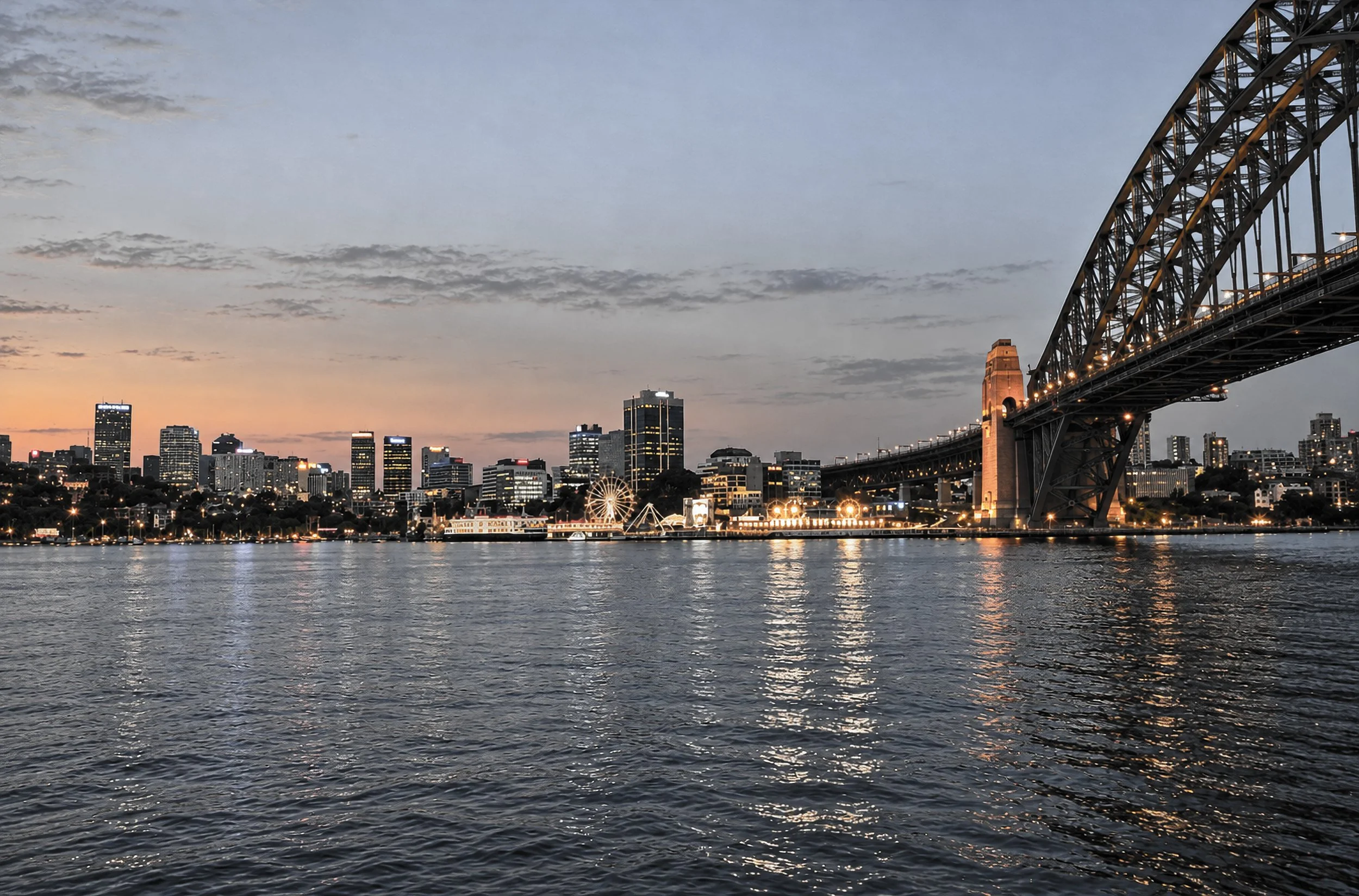 Sydney Harbour and Harbour Bridge - Sydney Harbour at dusk, viewed across the water towards the city skyline and Sydney Harbour Bridge. The evening lights of Circular Quay shimmer across the harbour, while the illuminated bridge dominates the skyline