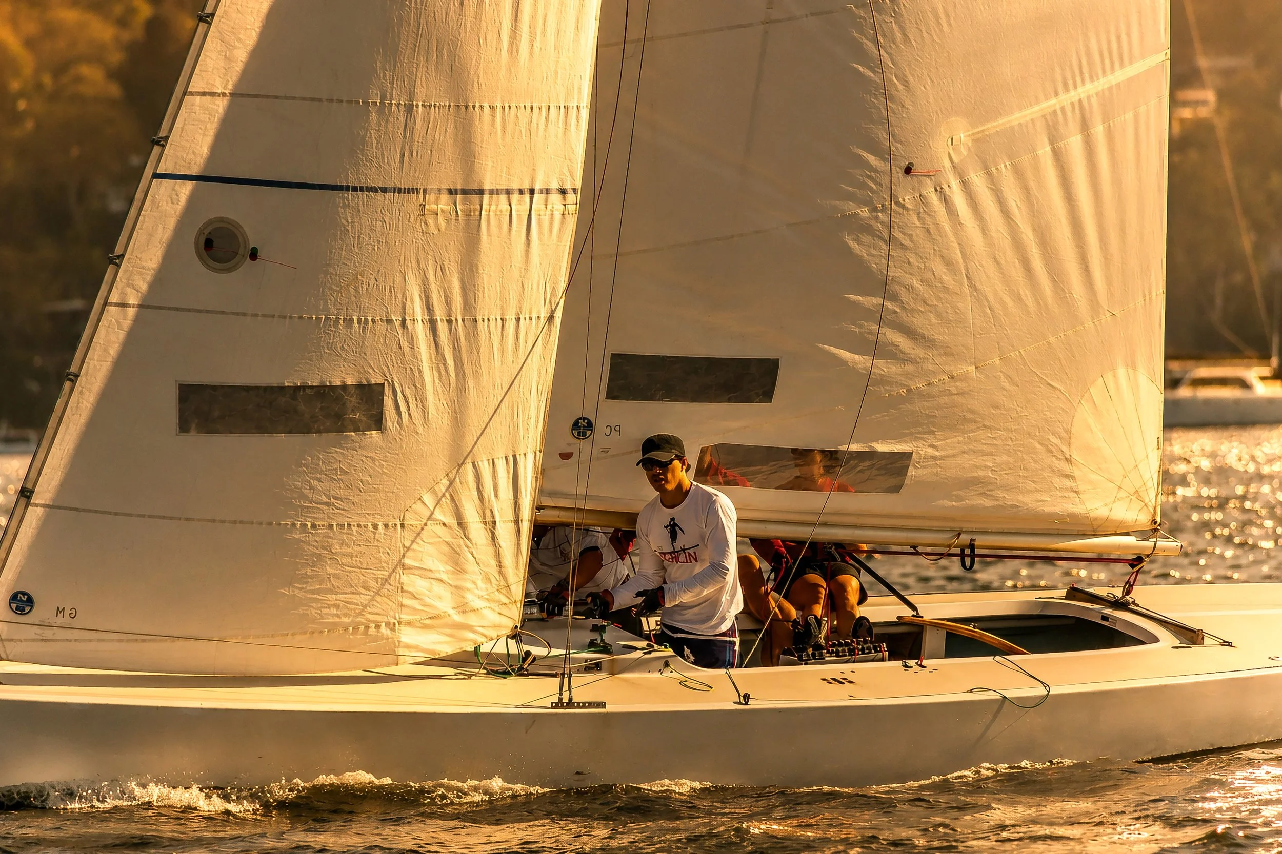 A sailboat with multiple crew members sailing on the water during sunset, with large sails catching the wind and the sunlight reflecting off the water.