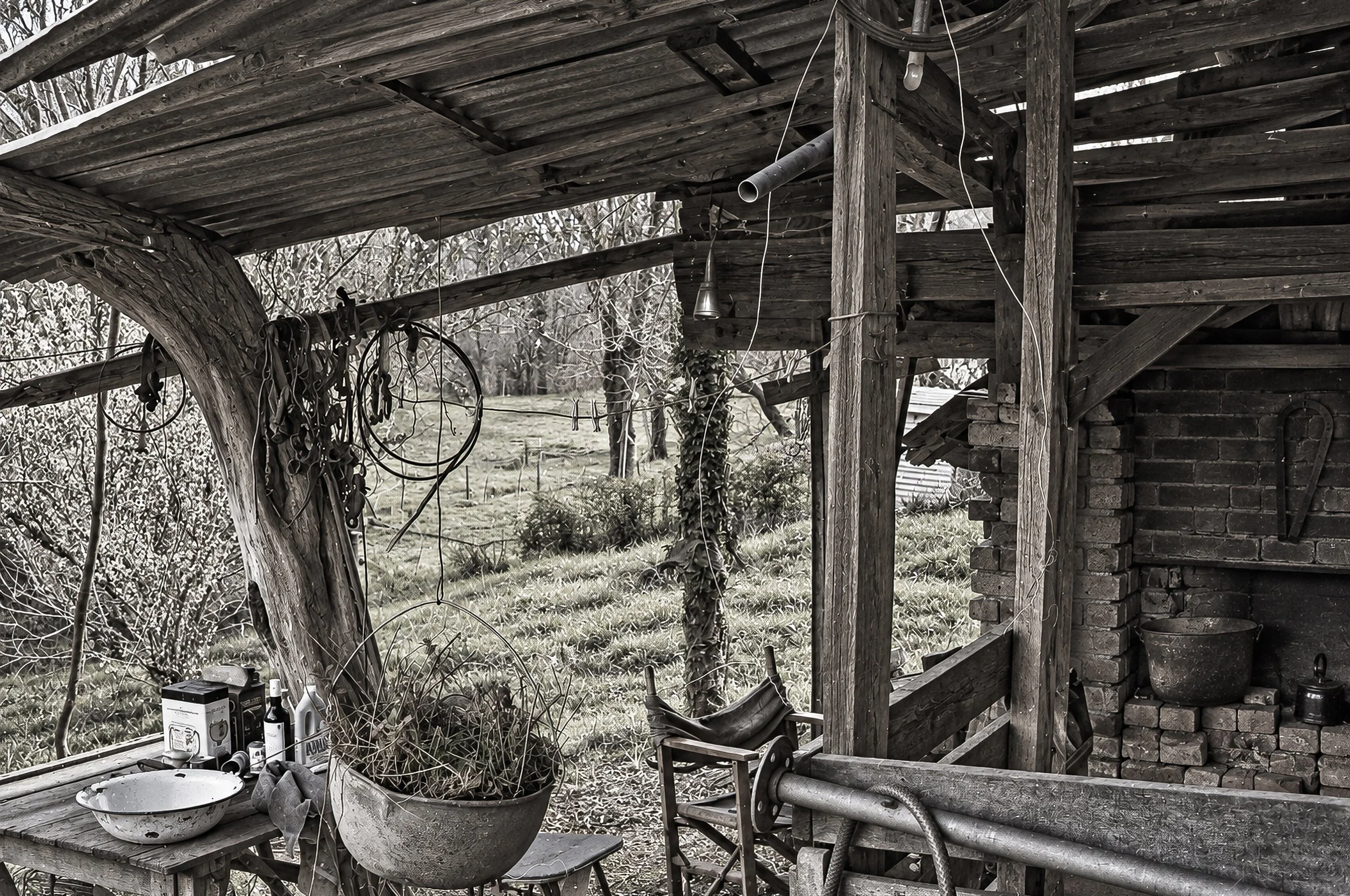 Life Under the Old Verandah - An old timber structure provides a sheltered outdoor living space surrounded by trees and bushland. The worn furniture, tools and objects left beneath the roof create an intimate portrait of rural life and the improvisat
