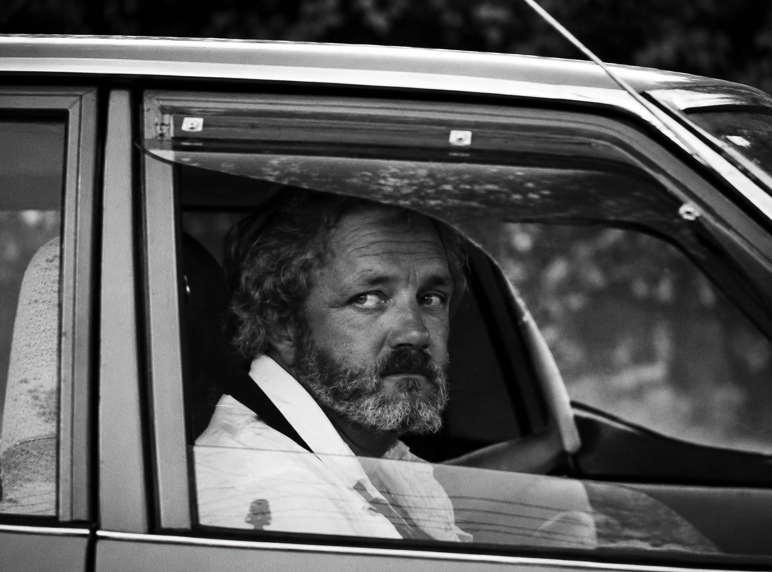 A Passing Look - A bearded man looks towards the camera from inside his car, his expression caught somewhere between curiosity and recognition. The window, reflections and dark interior reinforce the spontaneous, observational character of the photog