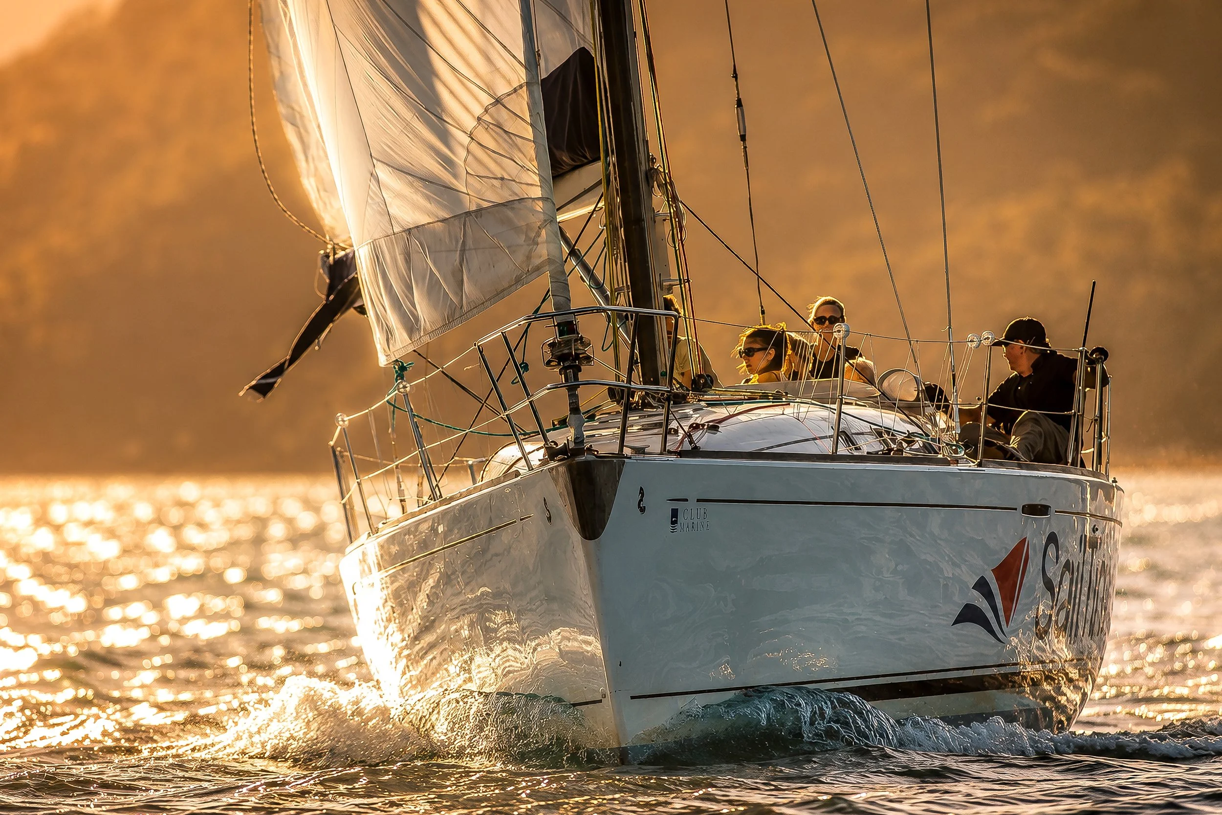 A sailboat on the water during sunset with three people aboard. The boat is moving forward, creating waves and reflections on the water, with a golden glow illuminating the scene.