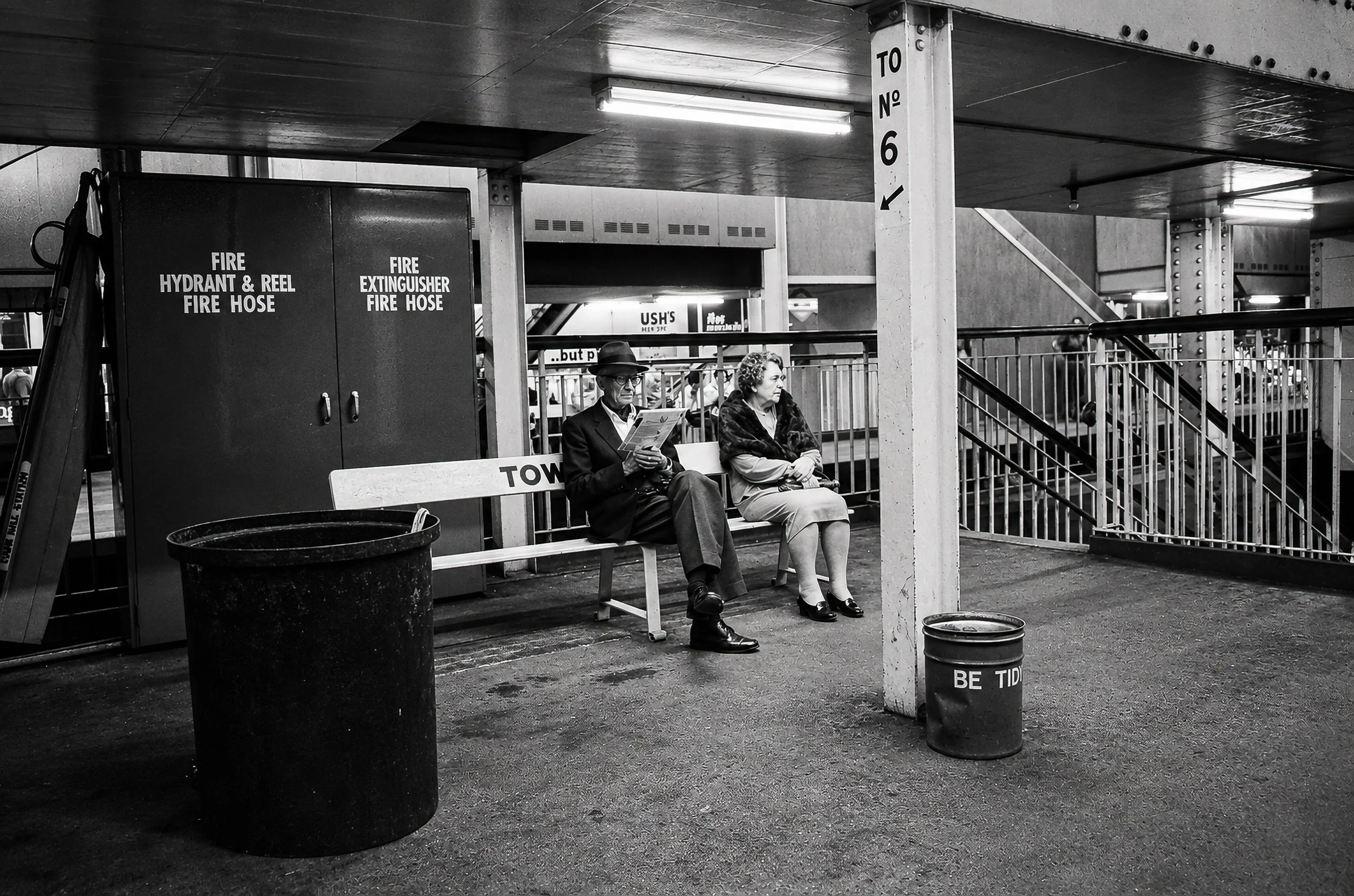 Waiting on the Platform - A quiet moment on a Sydney railway platform, with two passengers waiting on a bench beneath the station structure. The fire equipment, signs, fluorescent lighting and worn surfaces provide an authentic glimpse of the metropo